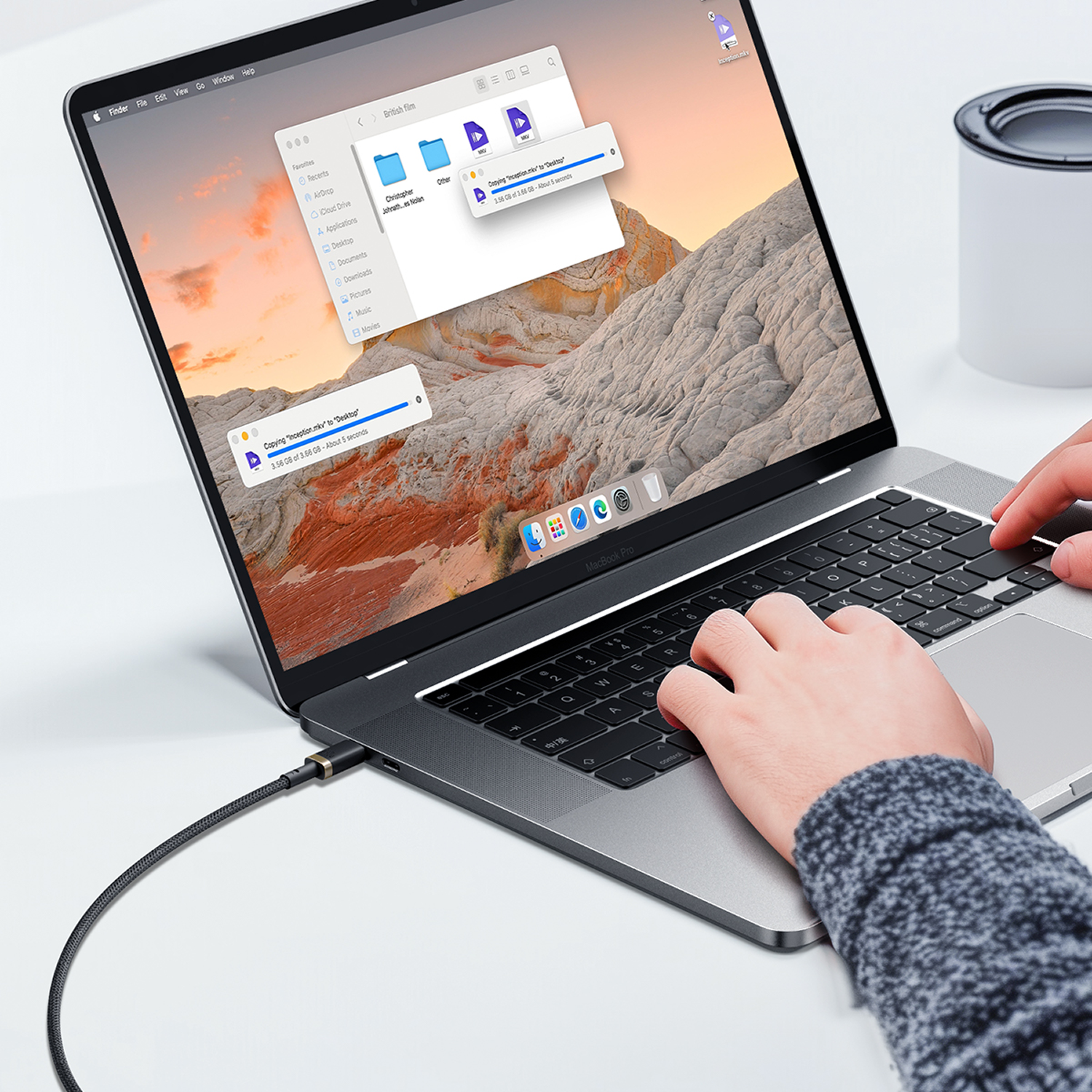 Una persona usa un MacBook Pro. La pantalla muestra una transferencia de archivos en curso. Una taza blanca es visible en el fondo.