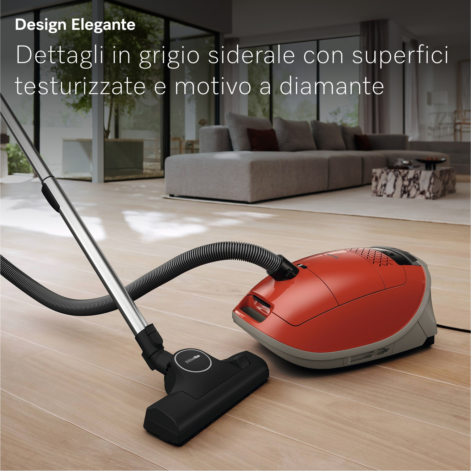 Un aspirapolvere rosso e grigio su un pavimento in legno, con tubo e spazzola neri. Un divano sullo sfondo.