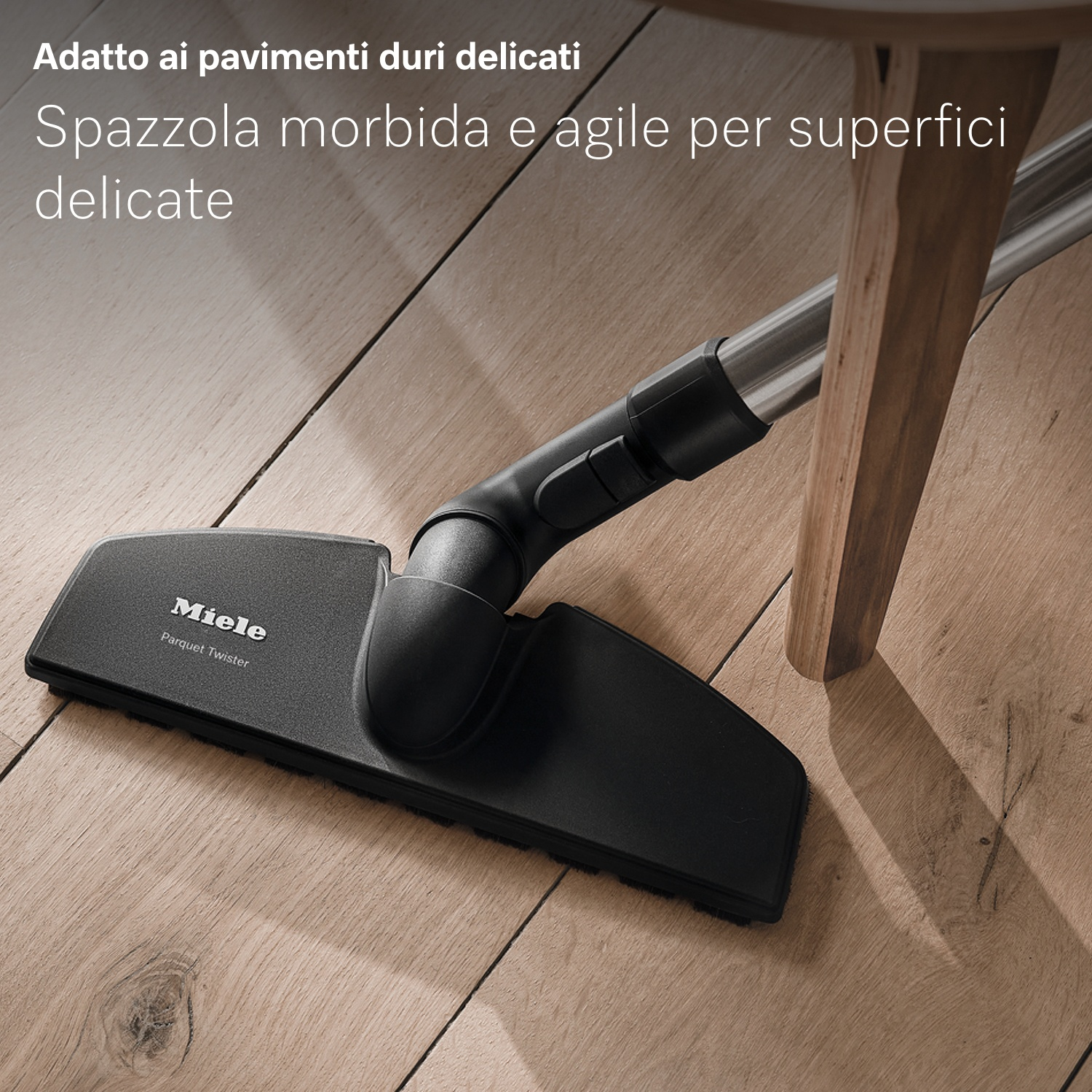 Testa aspirapolvere Miele nera su pavimento in legno. Piede del tavolo in legno a destra. Testo: 'Spazzola morbida e agile per superfici delicate'.