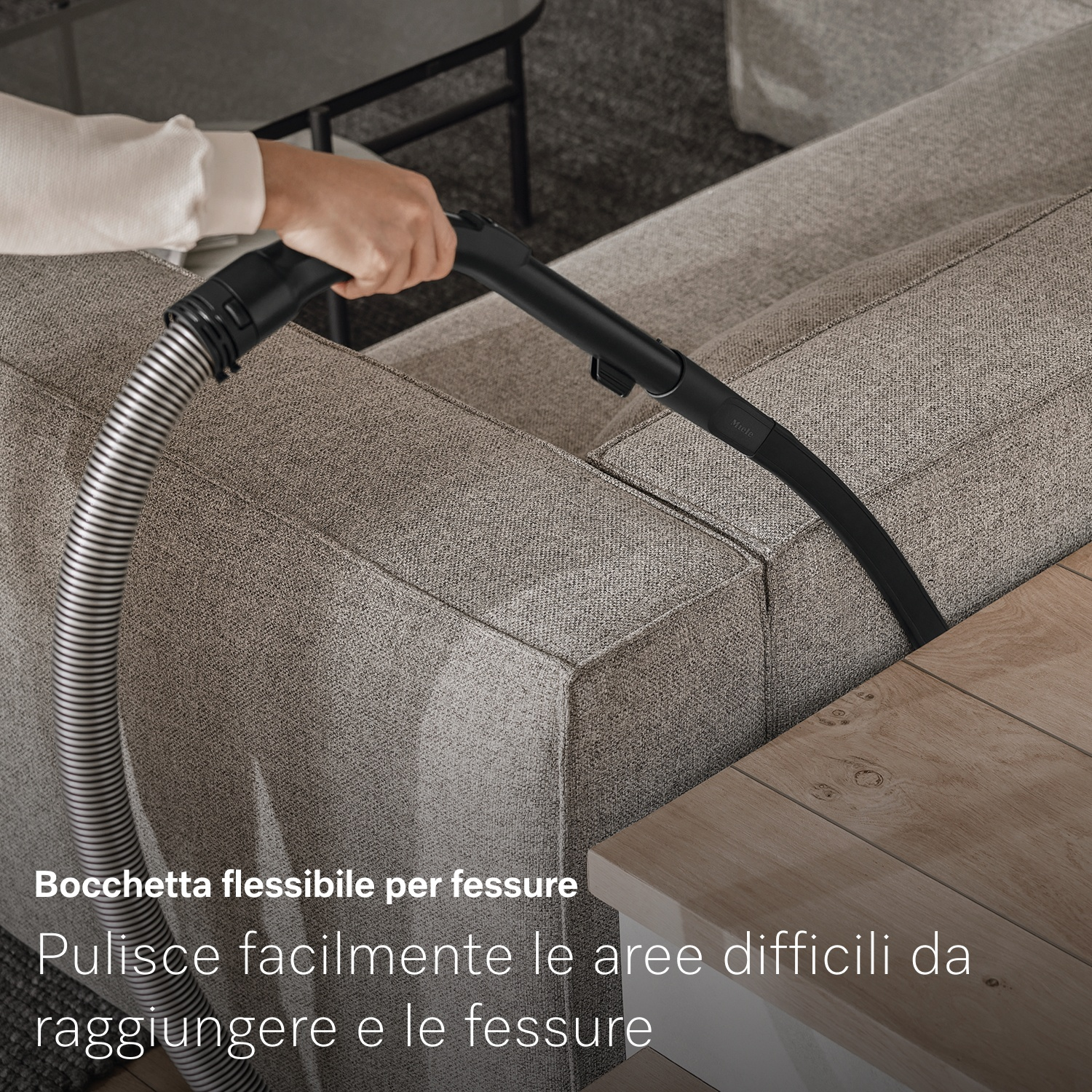 Una persona aspira la fessura di un divano con un accessorio nero. Un tavolo di legno è in primo piano. Il divano è grigio.