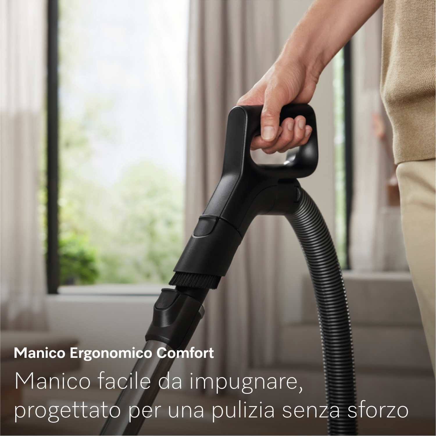Una persona tiene in mano l'impugnatura nera di un aspirapolvere. Lo sfondo mostra una finestra con vista sul verde e tende.