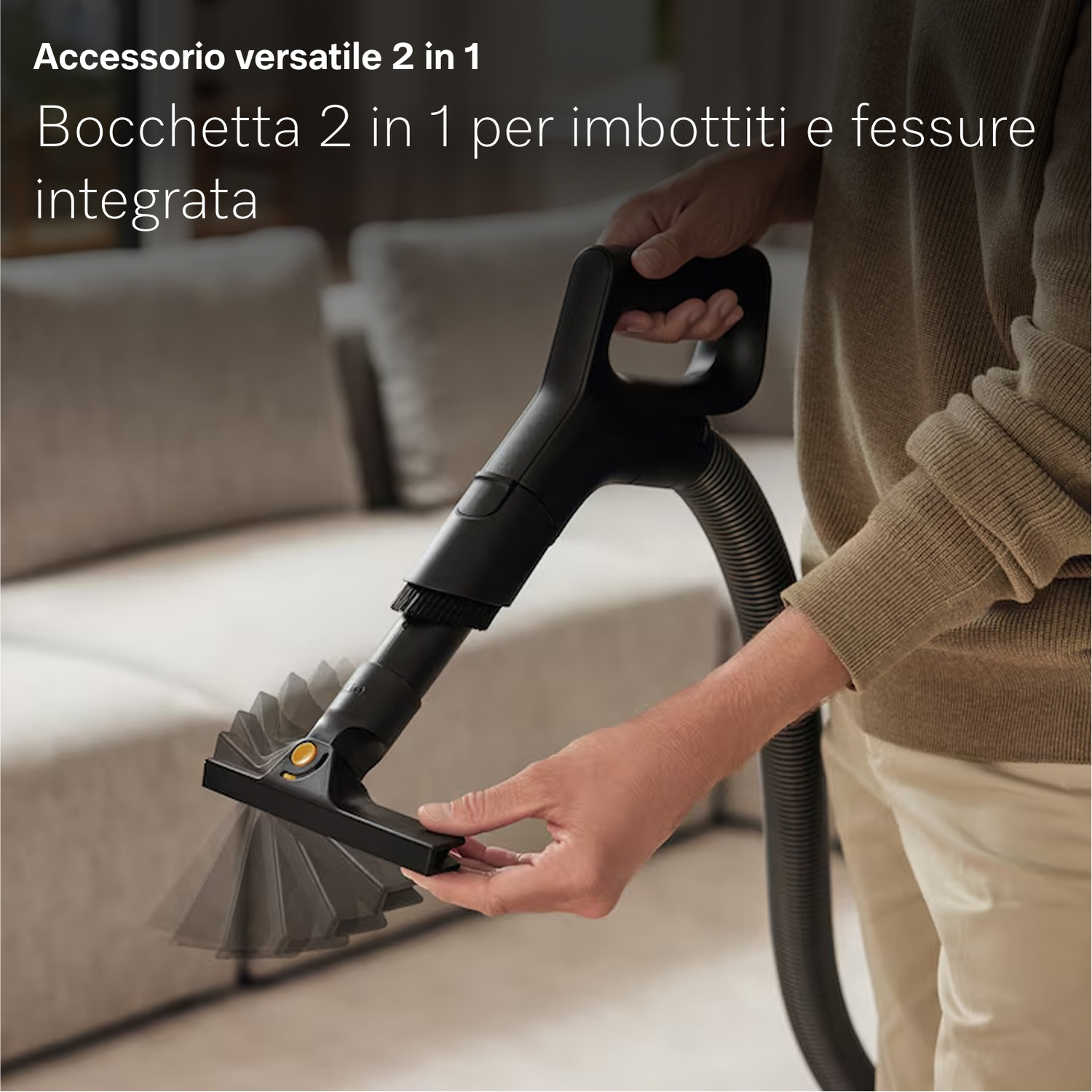 Una persona tiene in mano un accessorio nero per aspirapolvere. L'accessorio è vicino a un divano. La persona indossa un maglione e pantaloni beige.