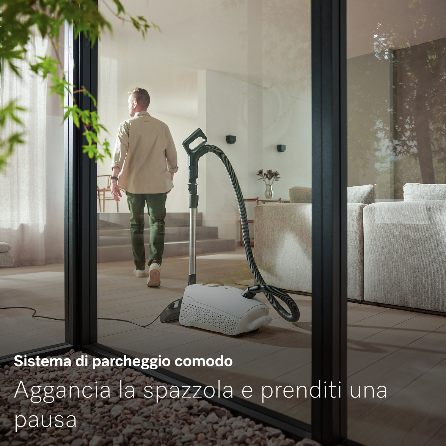 Un uomo si allontana. Un aspirapolvere in primo piano. Una finestra con porte a vetri mostra l'interno di una casa.