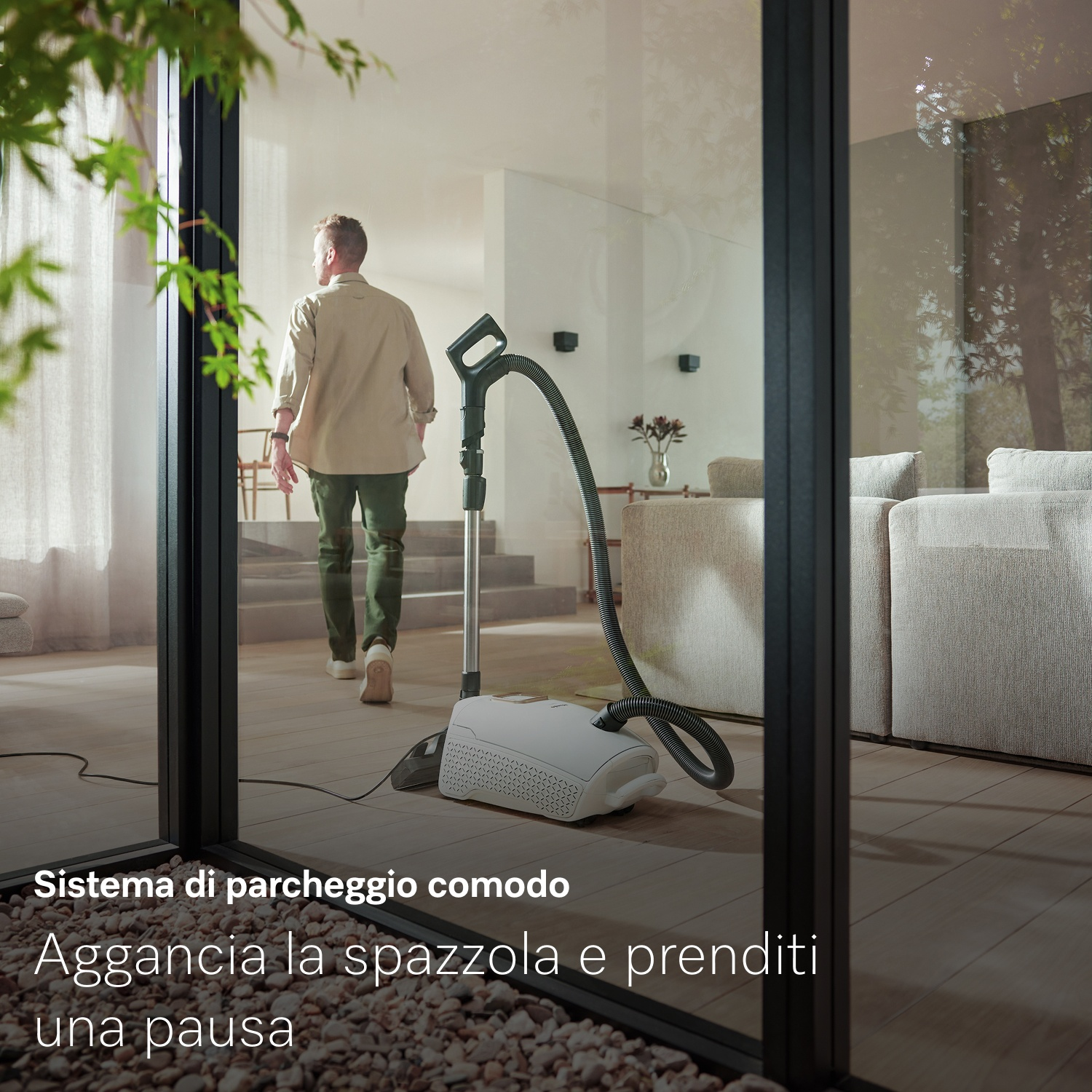 Un uomo si allontana da un aspirapolvere. Un moderno soggiorno è visibile attraverso una porta a vetri. Piante verdi in primo piano.