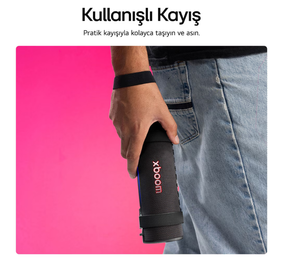 Bir kişi kırmızı 'xboom' yazılı siyah bir hoparlör tutuyor. Kişi kot pantolon ve siyah bir bileklik takıyor. Pembe arka plan.