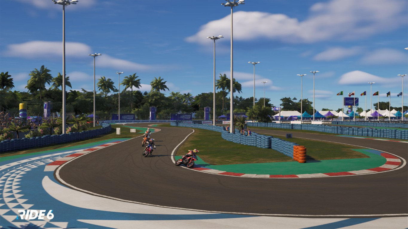 Motocykle ścigają się na torze z palmami, flagami i błękitnym niebem. Tor ma czerwono-białe obramowania.