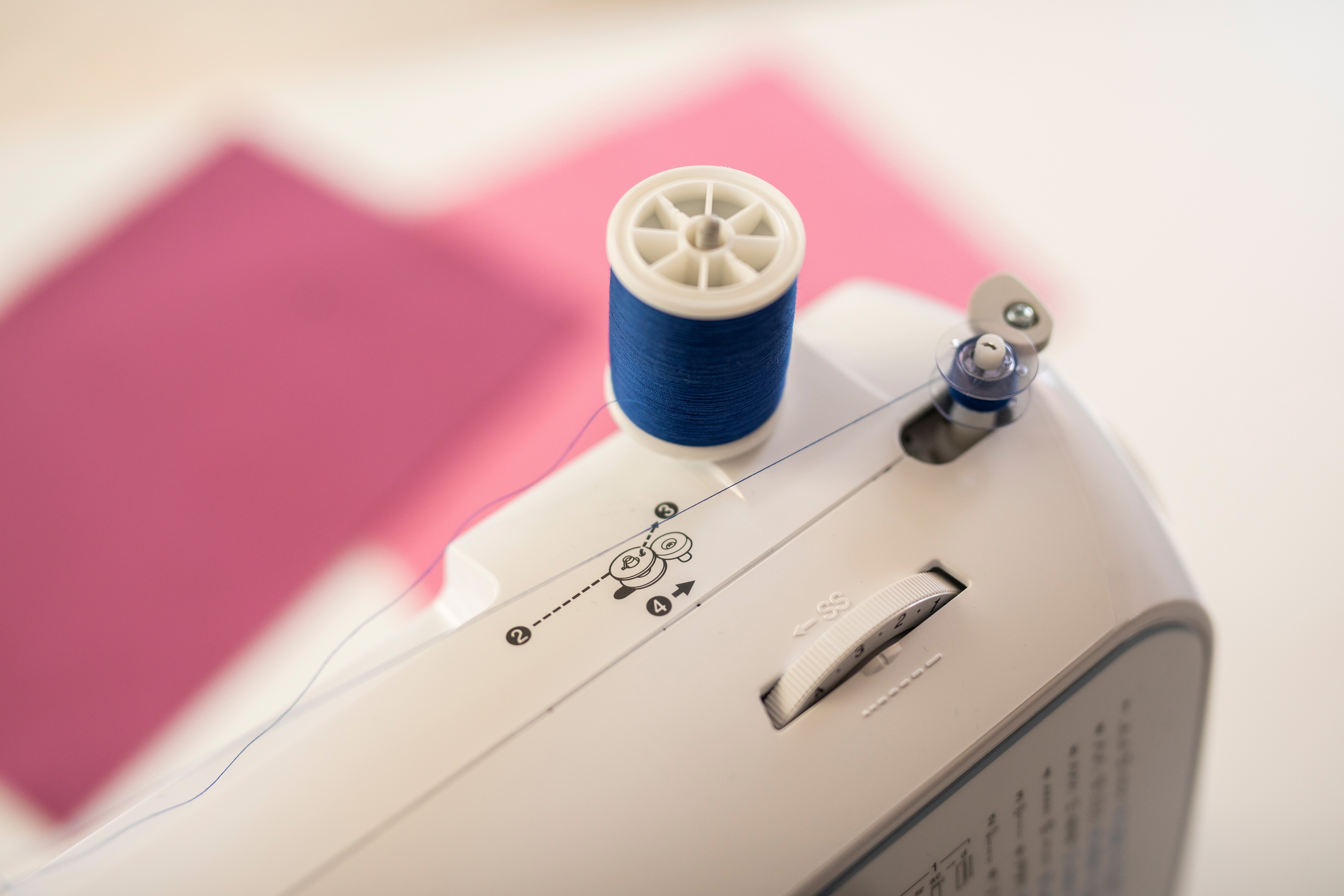 Nähmaschine mit blauer Garnspule. Rosa Stoff im Hintergrund, weißes Maschinengehäuse und ein Zifferblatt.