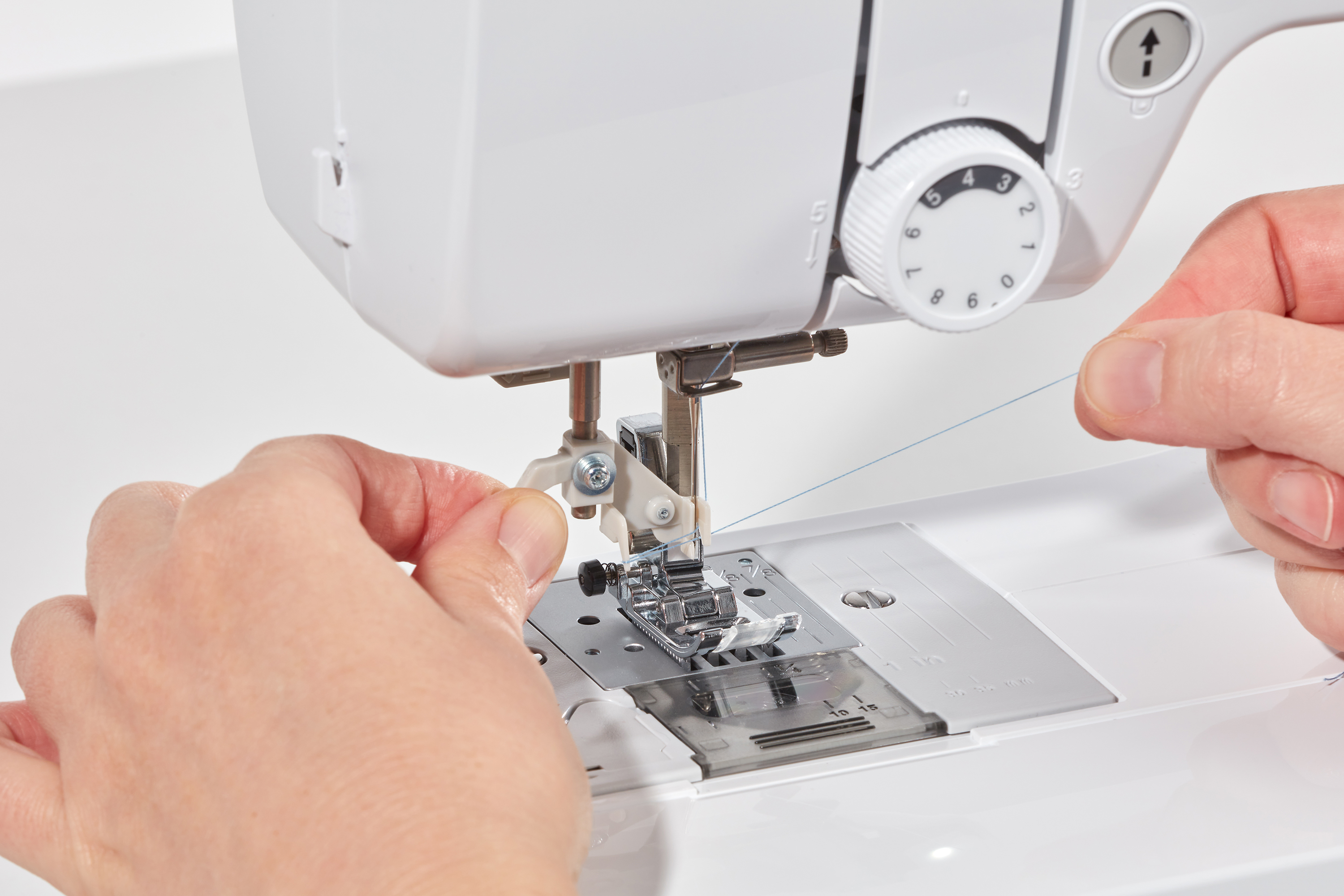 Hände, die eine Nähmaschine mit blauem Faden einfädeln. Weiße Maschine, Zifferblatt, Nadel und Platte sind sichtbar.