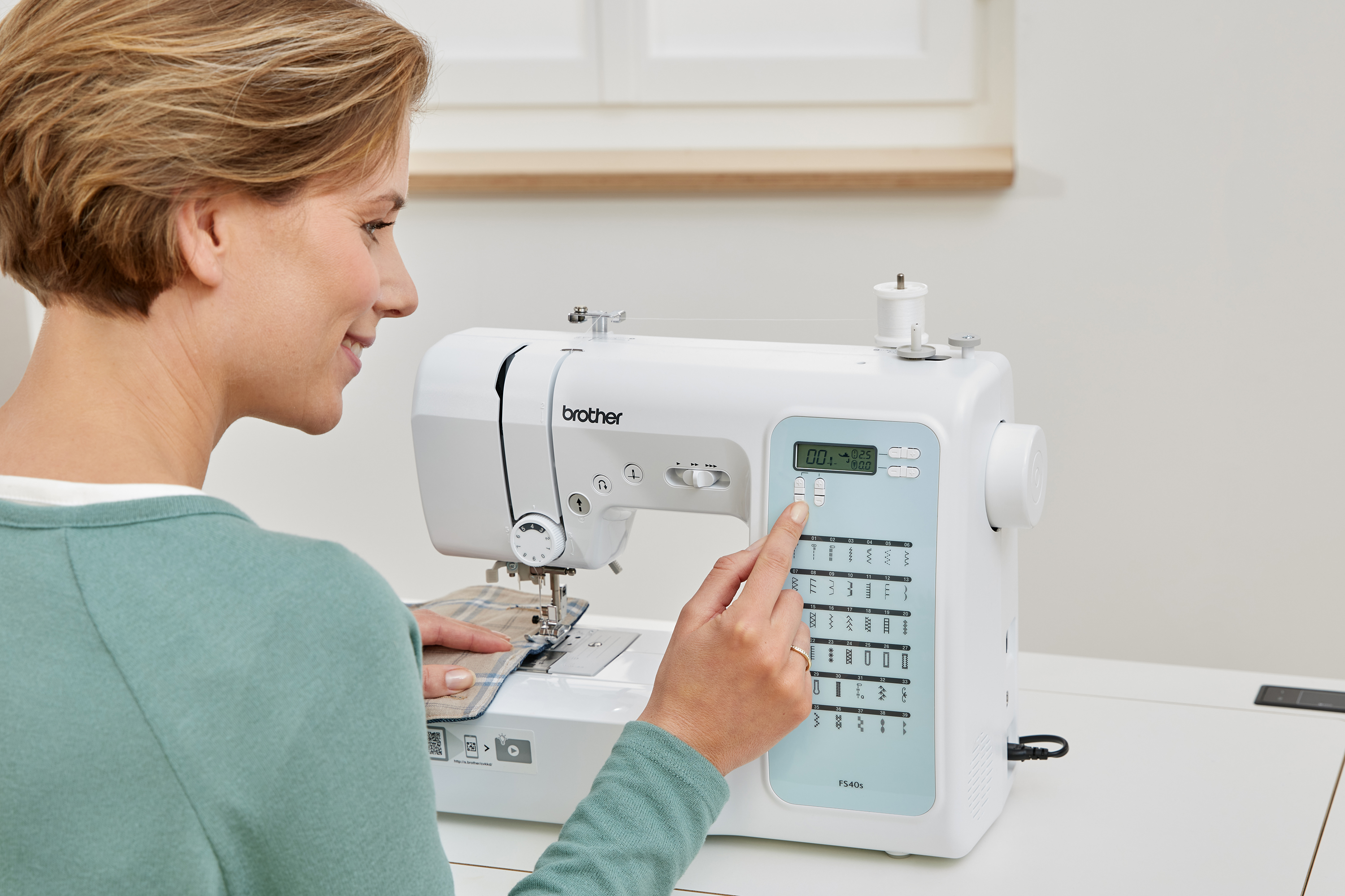 Eine Frau bedient eine weiße Brother-Nähmaschine. Sie näht karierten Stoff. Die Maschine hat eine Digitalanzeige.