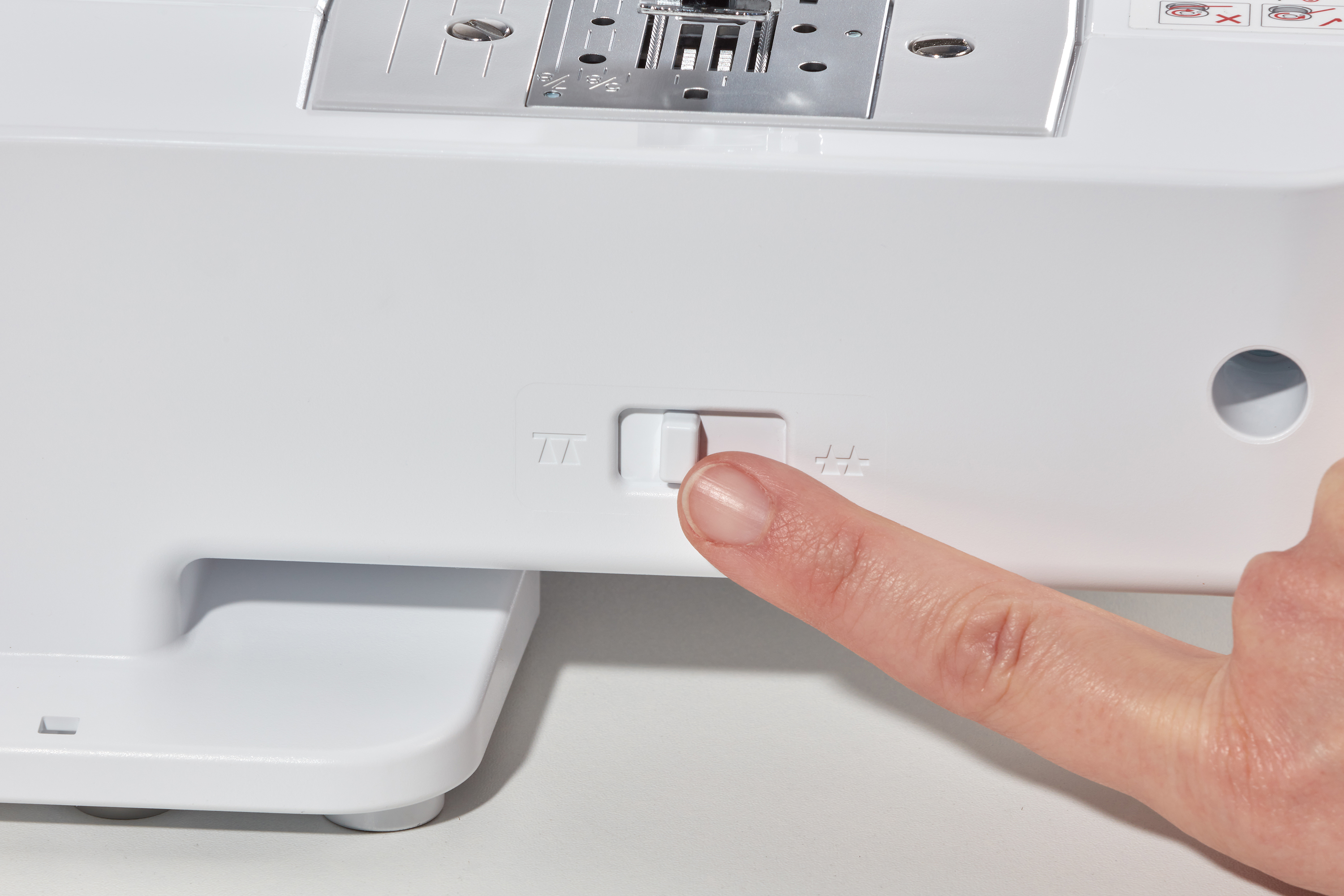 Ein Finger drückt einen weißen Schalter an einer weißen Nähmaschine. Der Schalter hat auf jeder Seite ein Symbol.