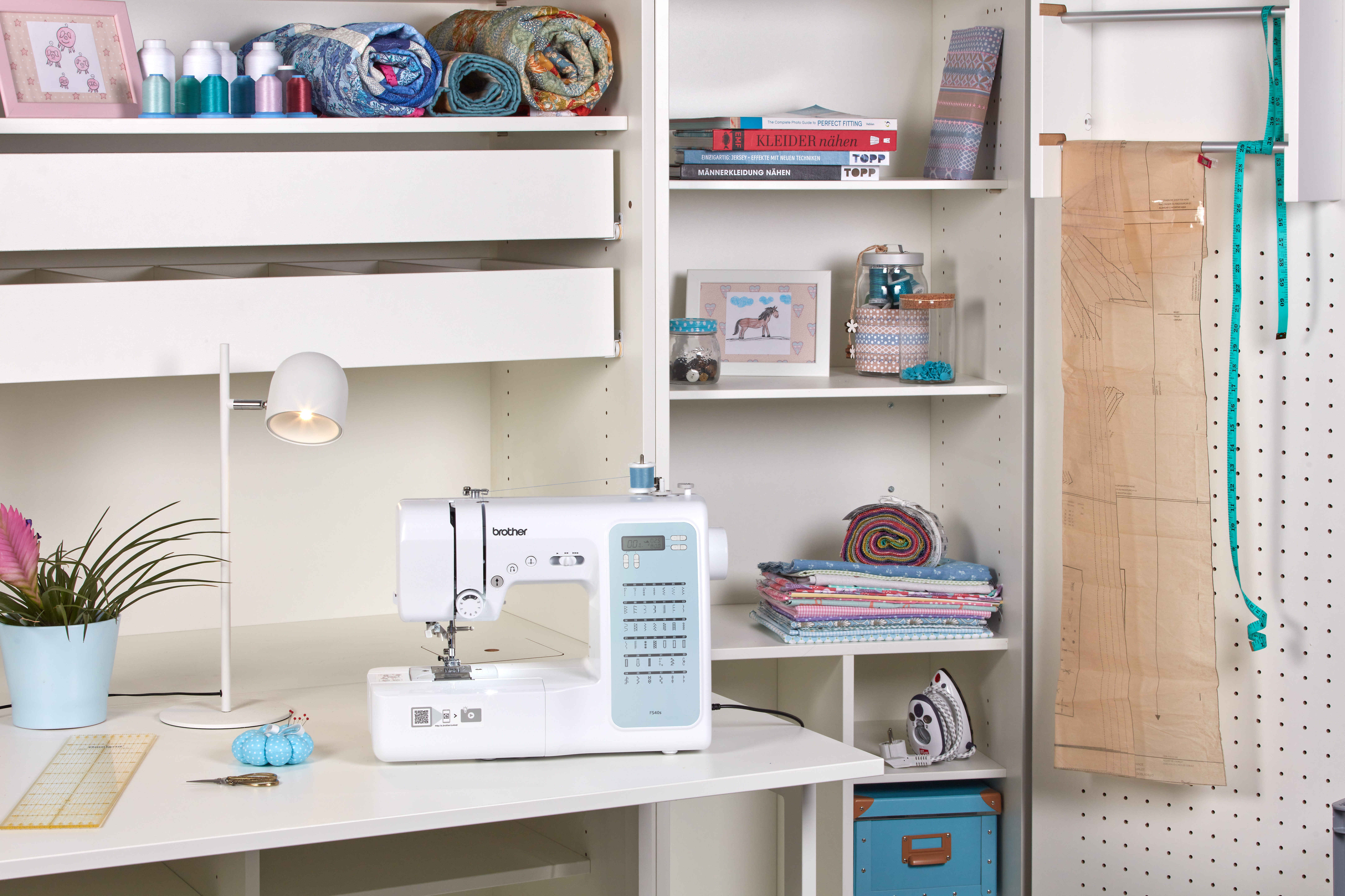 Nähzimmer mit weißer Maschine, Stoffrollen, Regalen mit Nähzubehör und einer Pflanze. Ein Schnittmuster hängt an der Wand.