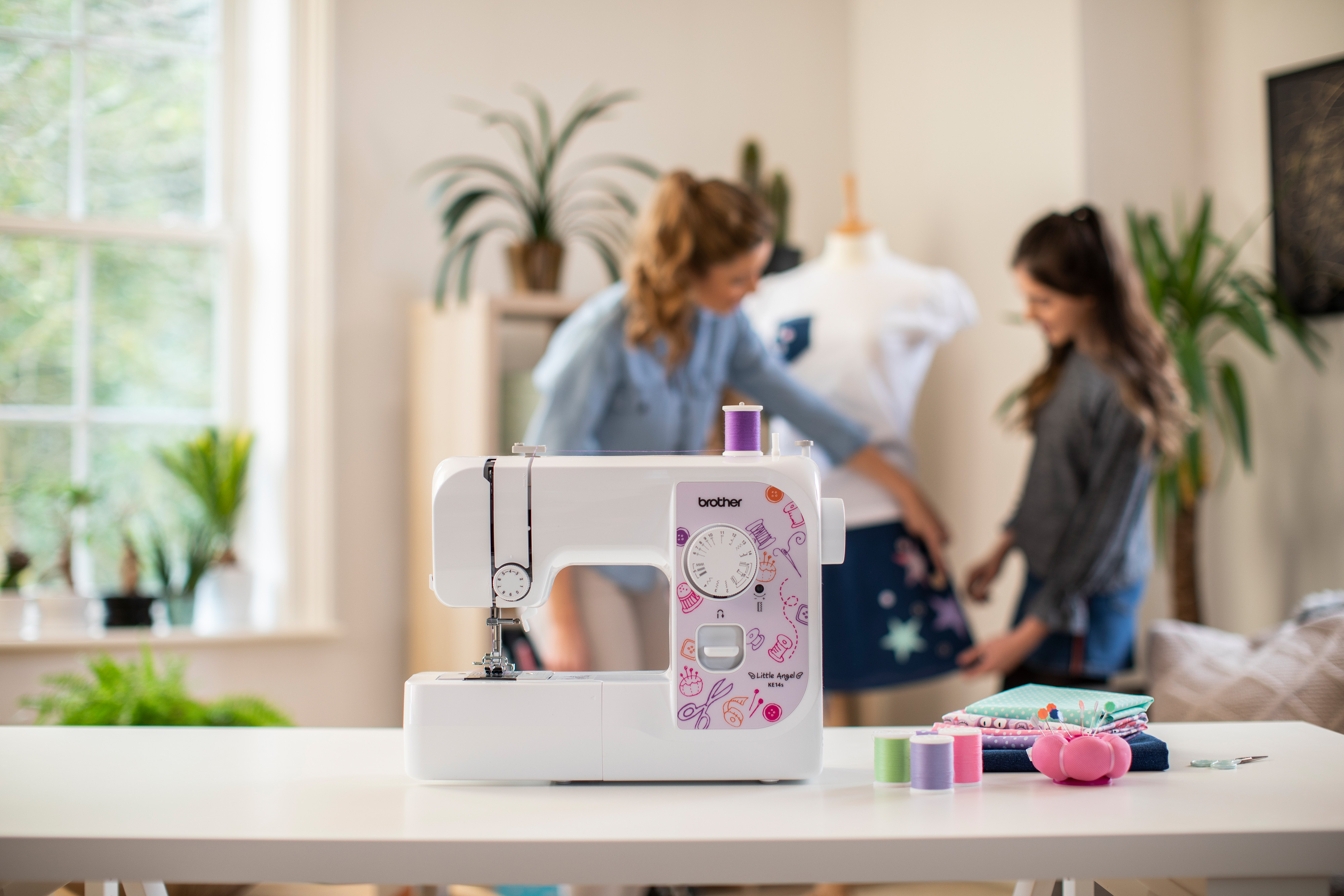 Eine Nähmaschine auf einem weißen Tisch. Zwei Frauen arbeiten mit Stoff. Im Hintergrund sind ein Fenster und Pflanzen zu sehen.