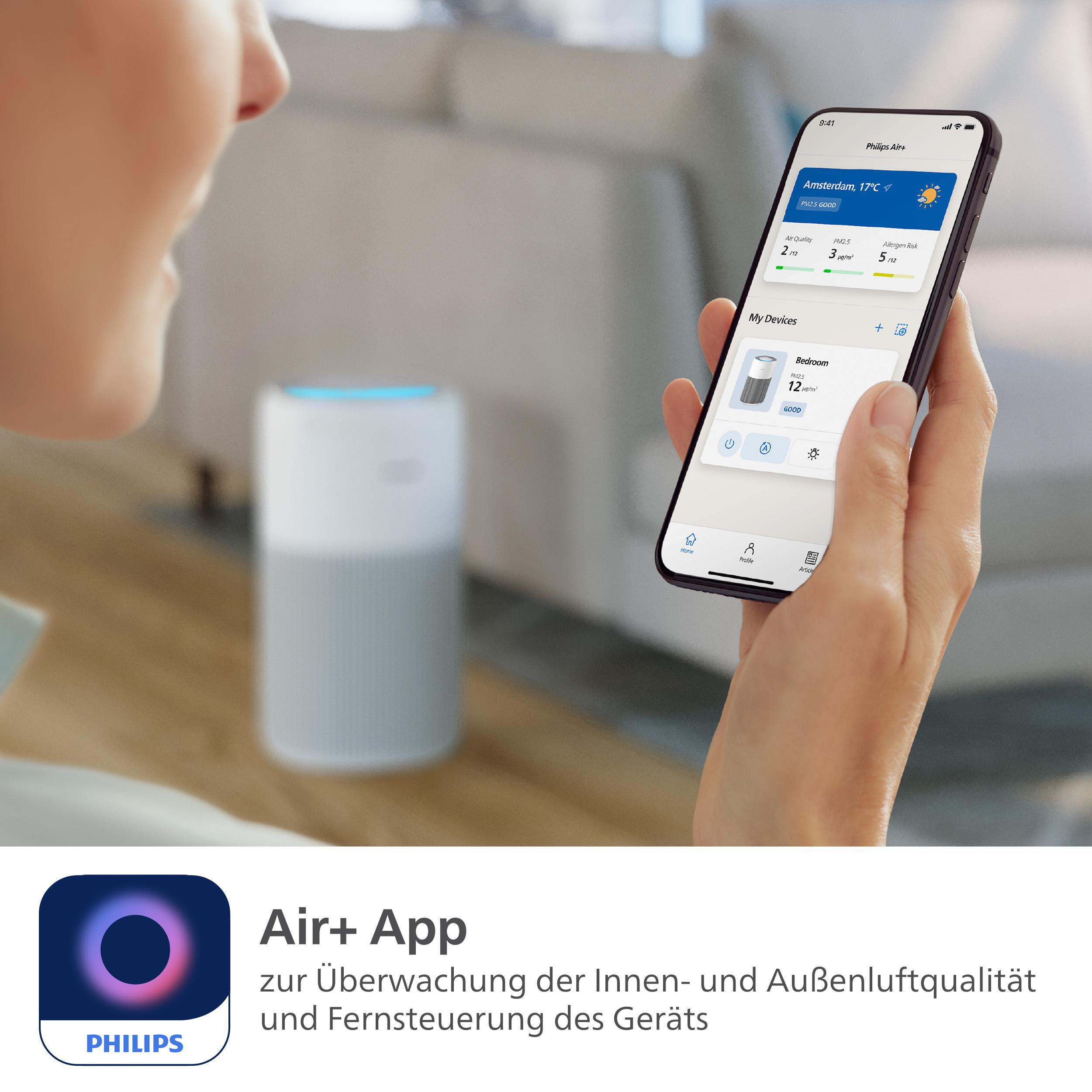 Eine Person hält ein Telefon mit einer Luftqualitäts-App. Im Hintergrund befindet sich ein weißer Luftreiniger.