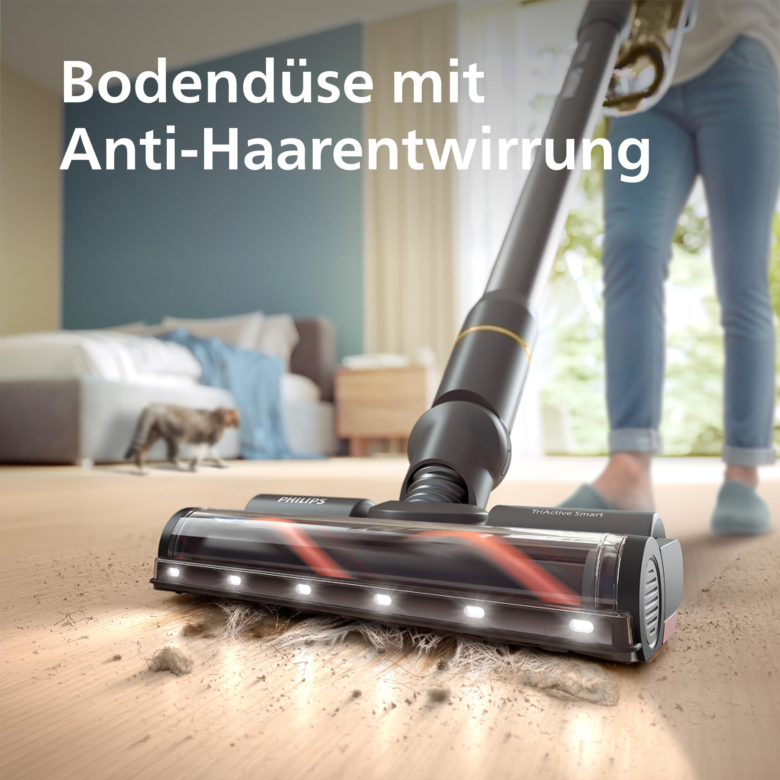 Eine Person benutzt einen Staubsauger. Der Staubsauger steht auf einem Holzboden, mit einer Katze und Möbeln im Hintergrund.