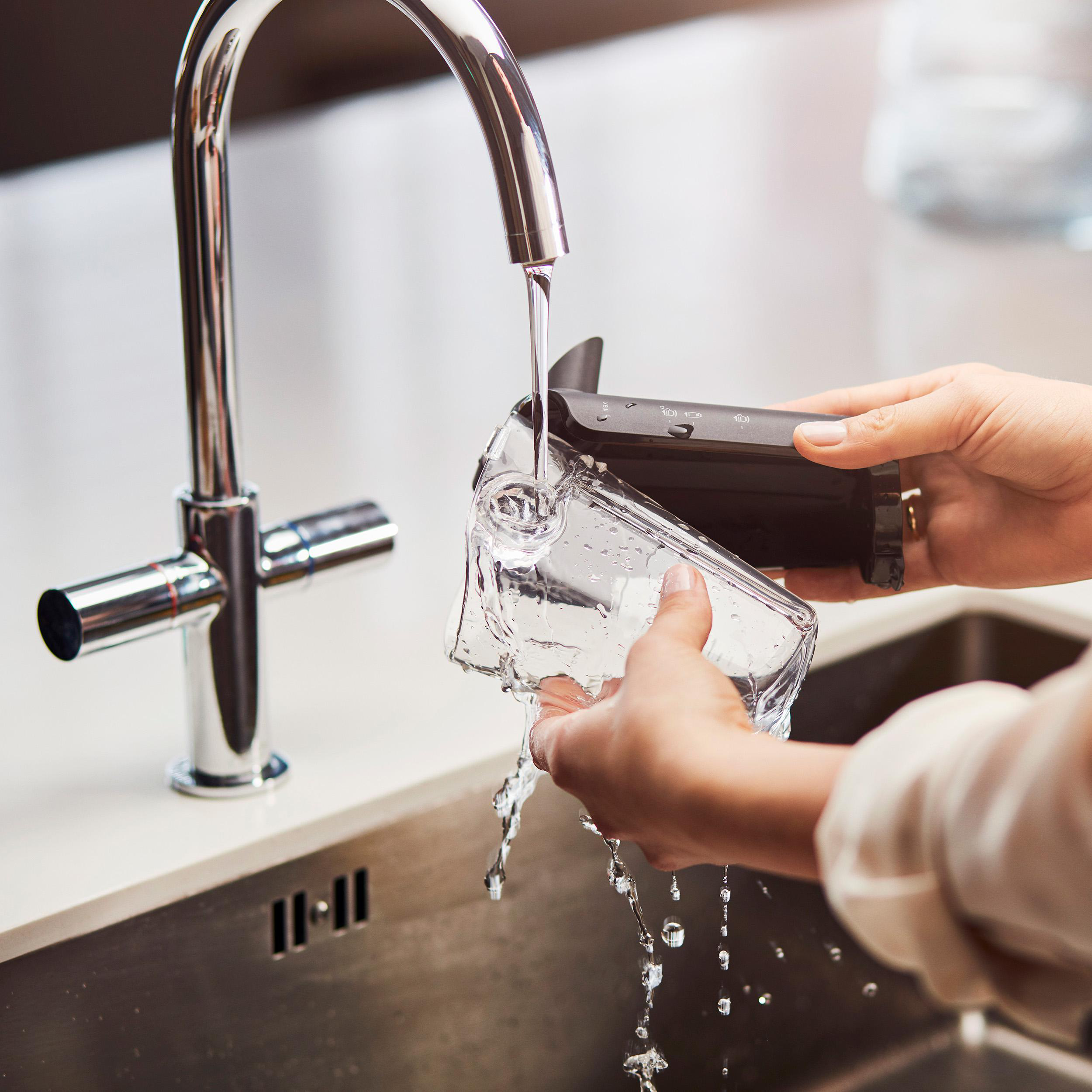 Hände waschen einen durchsichtigen Glasbehälter unter einem Chromhahn. Wasser fließt in den Behälter, über ein Waschbecken.