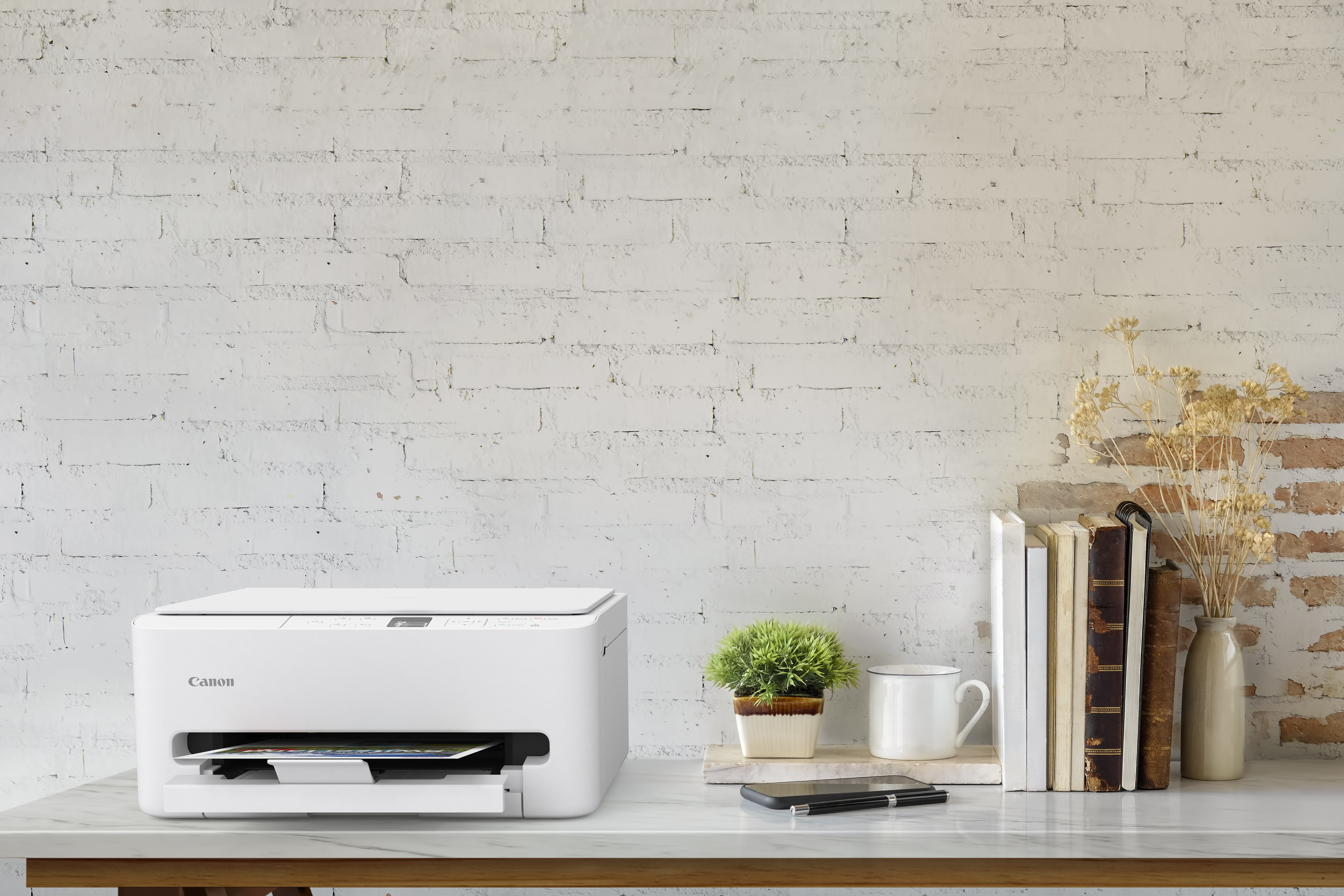 Biała drukarka Canon stoi na marmurowym biurku. Książki, roślina i kubek znajdują się na biurku na tle białej ceglanej ściany.