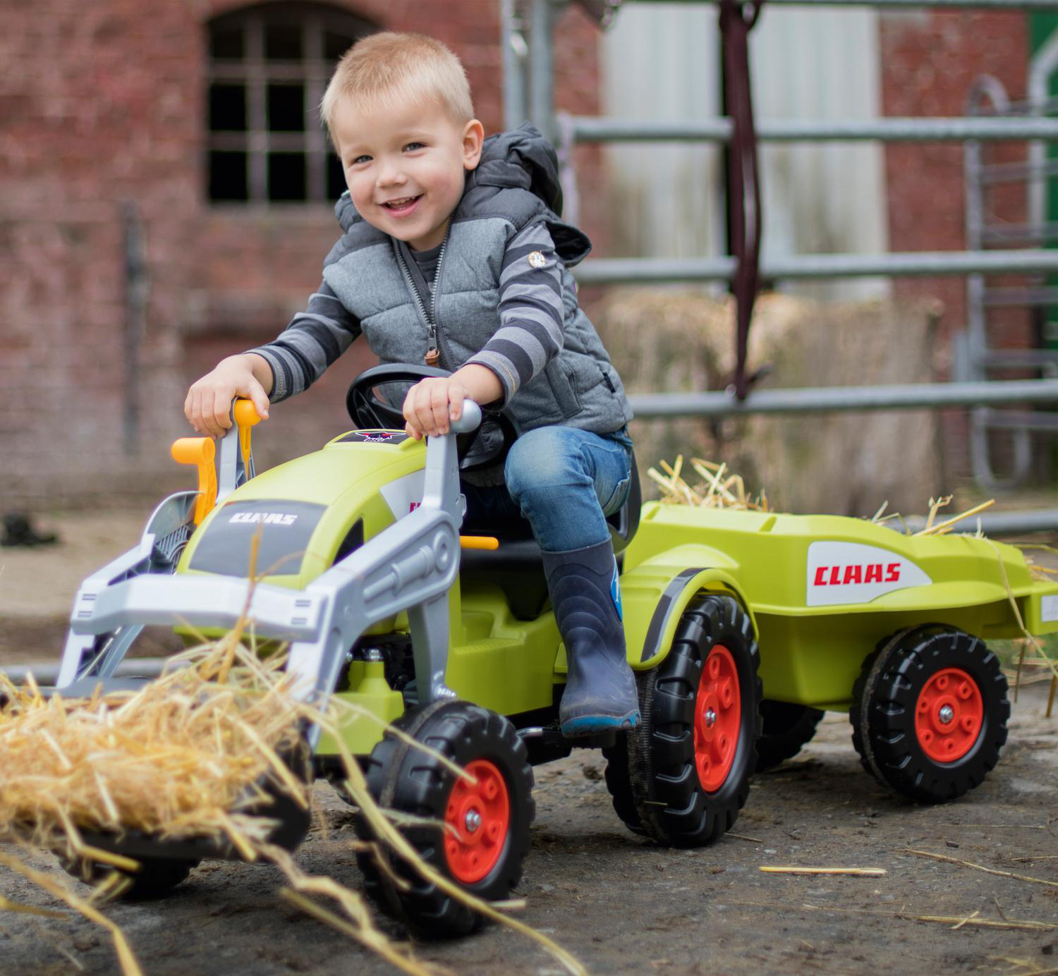 Ein lächelnder Junge in einer Weste fährt einen Claas-Spielzeugtraktor mit Anhänger. Der Traktor ist grün, mit Heu. Im Hintergrund ist ein Backsteingebäude.
