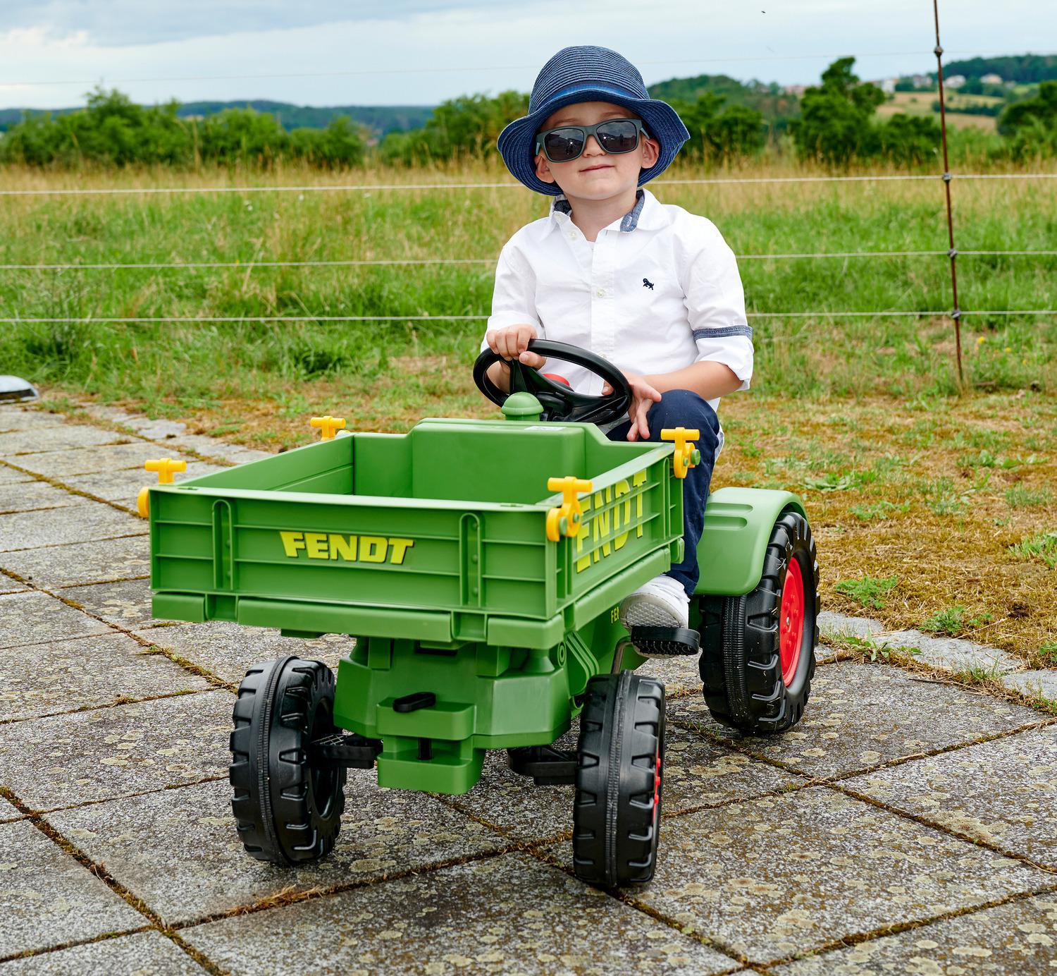 Ein Junge mit Sonnenbrille und blauem Hut sitzt auf einem grünen Spielzeugtraktor. Der Hintergrund ist eine Wiese und ein bewölkter Himmel.