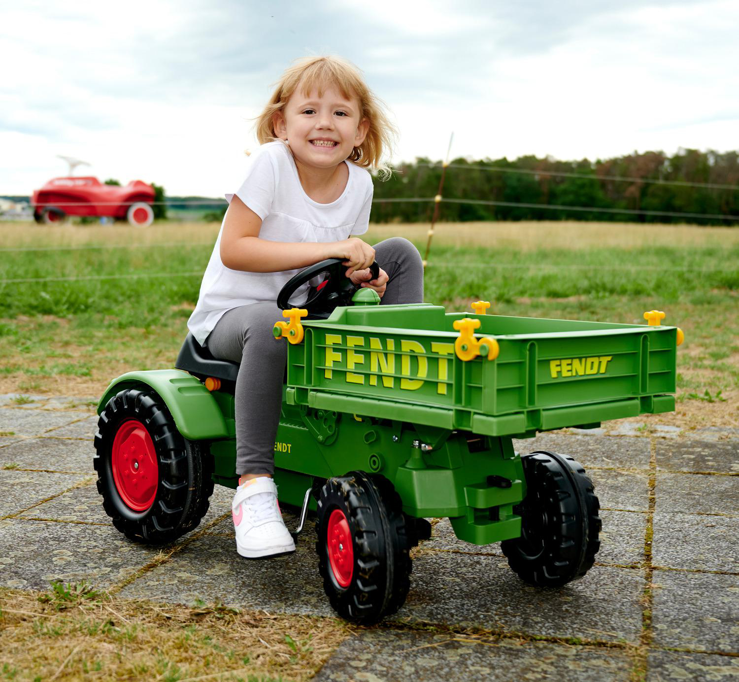 Ein Mädchen sitzt lächelnd auf einem grünen Fendt-Traktorspielzeug. Ein rotes Autospielzeug ist im Hintergrund auf einem Feld.
