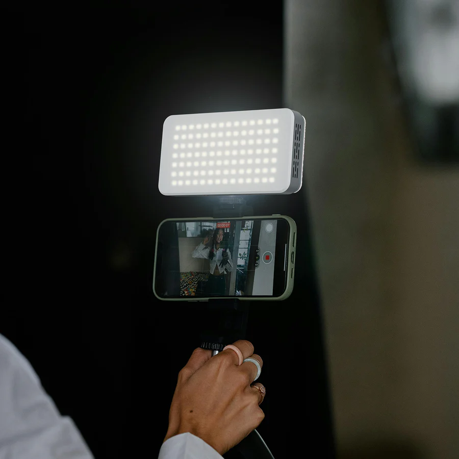 Une personne tenant un téléphone enregistrant une vidéo avec une lumière. Le téléphone est dans un étui vert. La personne porte des bagues.