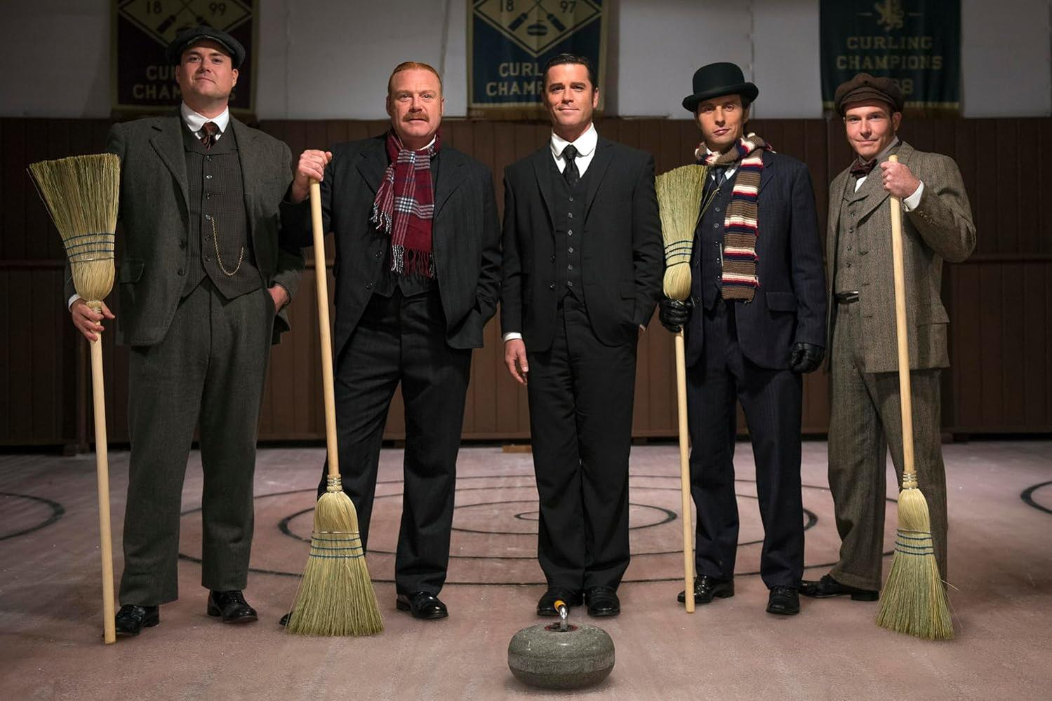 Fünf Männer in Vintage-Kleidung halten Besen und posieren in einer Curling-Arena. Vorne steht ein Curling-Stein. Im Hintergrund hängen Banner.