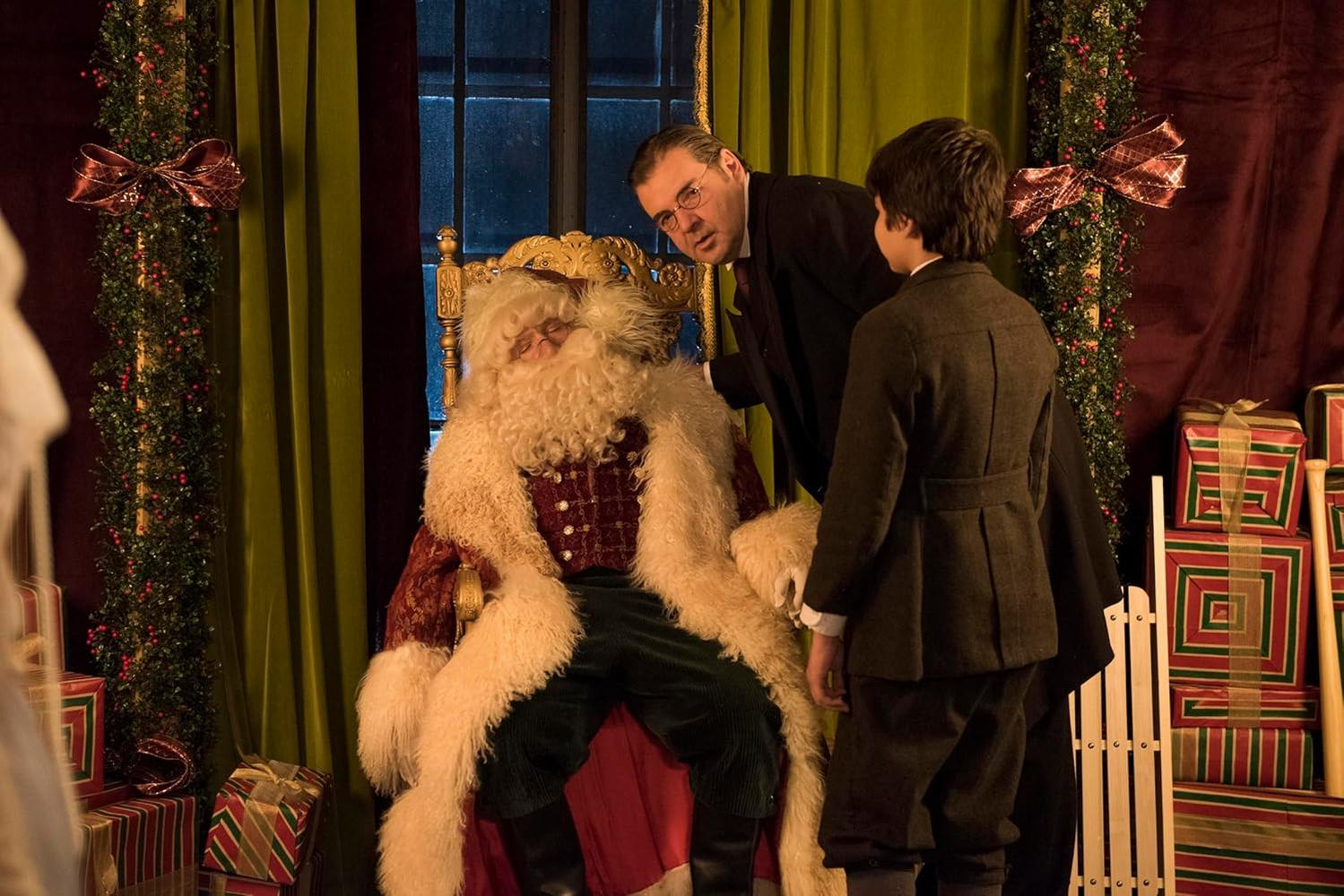 Der Weihnachtsmann sitzt, flankiert von zwei Männern, in einem dekorierten Raum. Geschenke und festliches Grün sind sichtbar.