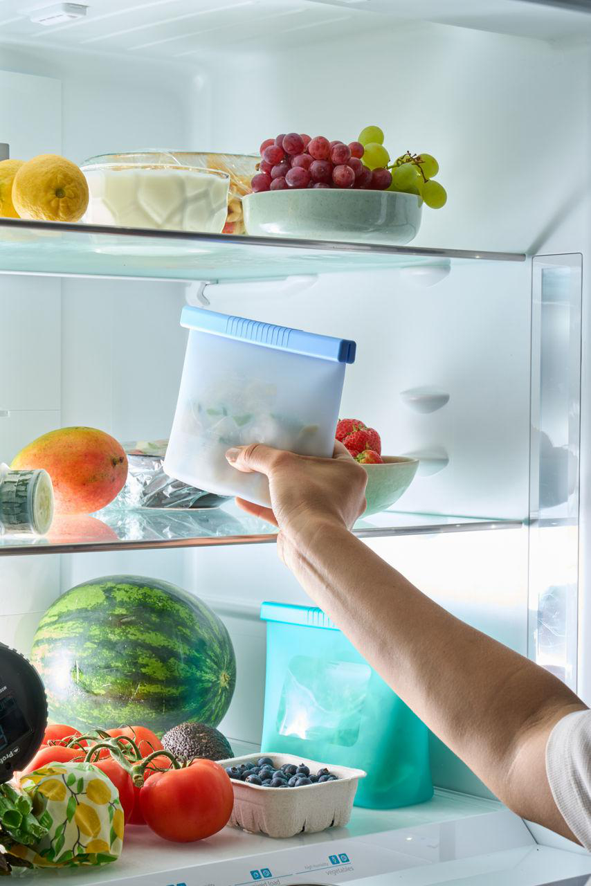 Eine Hand hält eine Tüte in einem Kühlschrank voller Obst und Gemüse. Trauben, Mangos, Tomaten und Wassermelone sind sichtbar.
