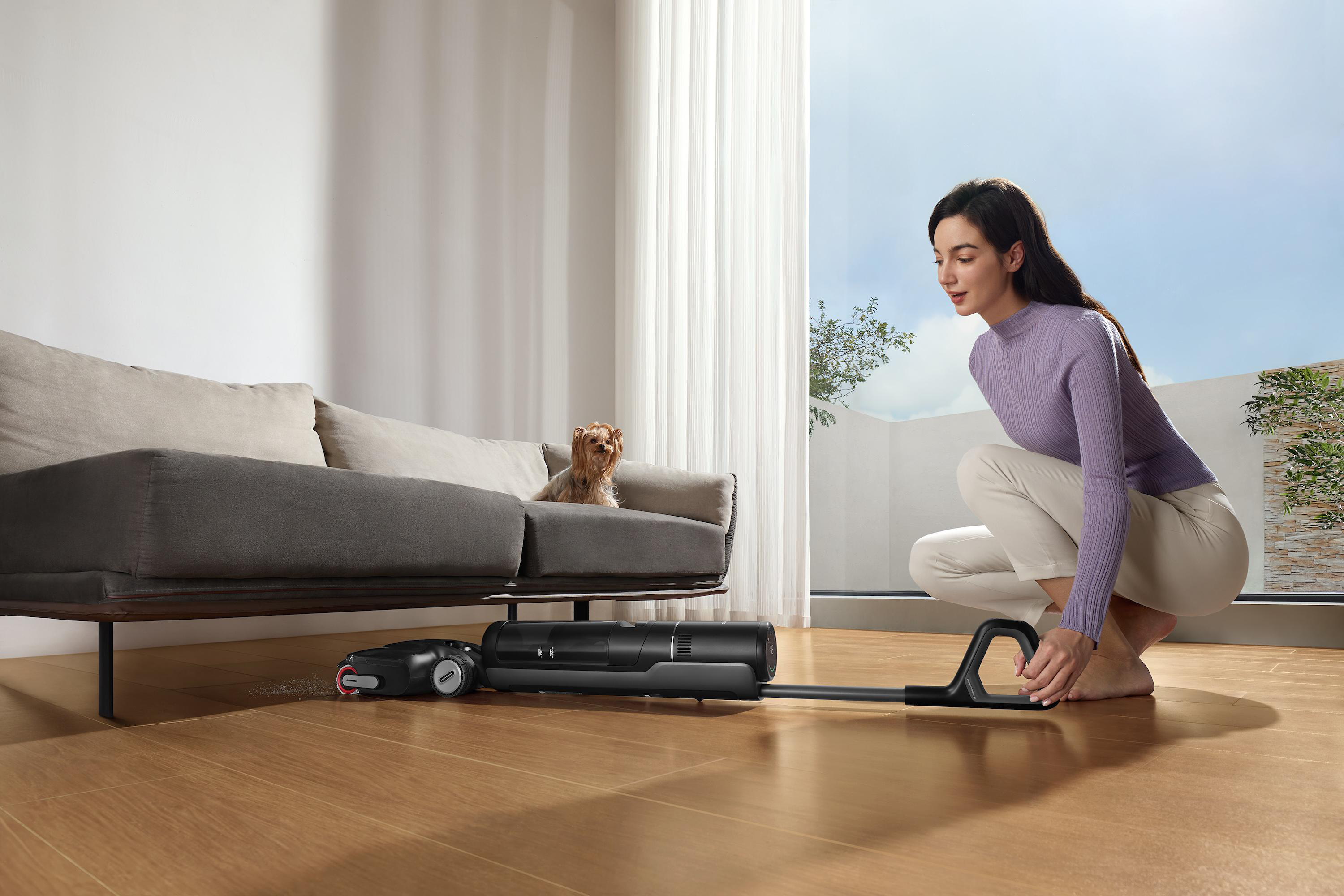 Frau mit Staubsauger, Hund auf Couch. Helles Zimmer, Holzboden. Himmel durch Fenster sichtbar.