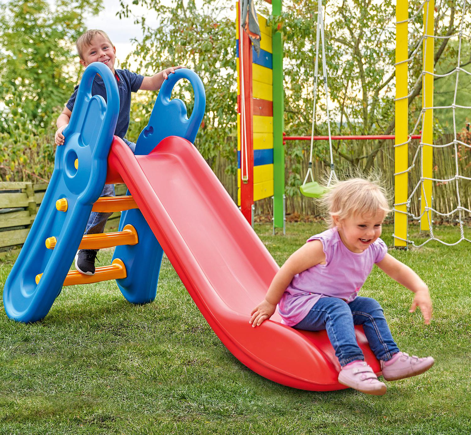 Zwei Kinder spielen auf einer rot-blauen Rutsche im Garten. Eines klettert, das andere rutscht runter. Spielplatz im Hintergrund.