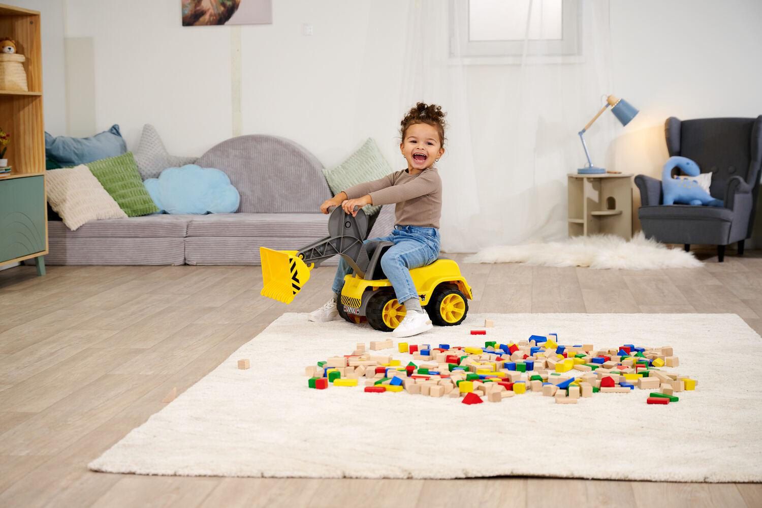 Ein lächelndes Kind auf einem gelben Spielzeugbagger, umgeben von Blöcken auf einem weißen Teppich. Das Zimmer hat ein Sofa und andere Spielzeuge.