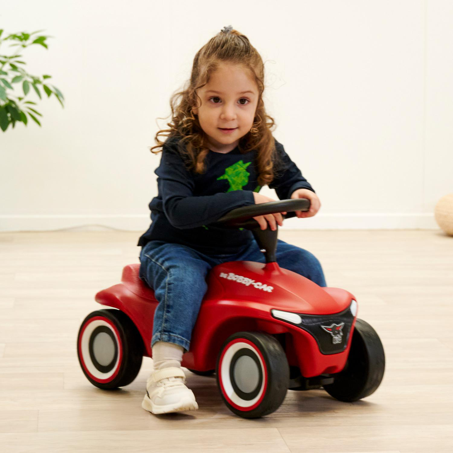 Ein junges Mädchen sitzt in einem roten Spielzeugauto und blickt in die Kamera. Sie trägt ein dunkelblaues Hemd und Jeans. Das Auto steht auf einem Holzboden.