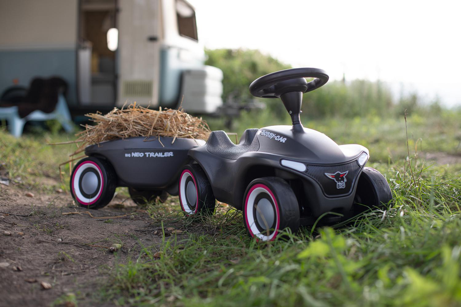 Ein schwarzes Spielzeugauto mit Anhänger voller Heu steht auf Gras. Im Hintergrund ist ein Wohnmobil zu sehen.