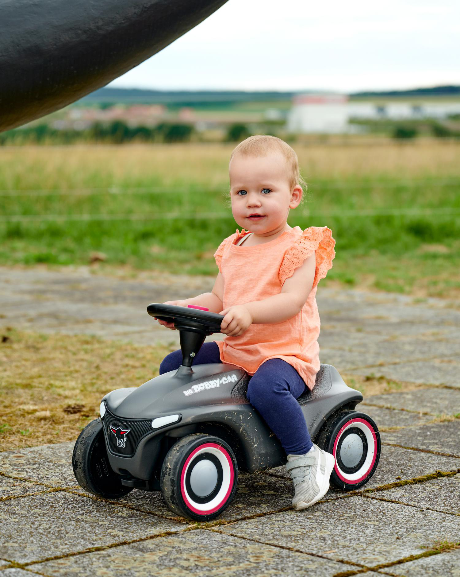 Ein Baby sitzt draußen auf einem grauen Spielzeugauto. Das Baby trägt ein orangefarbenes Hemd und blaue Hosen. Im Hintergrund befinden sich grüne Felder.