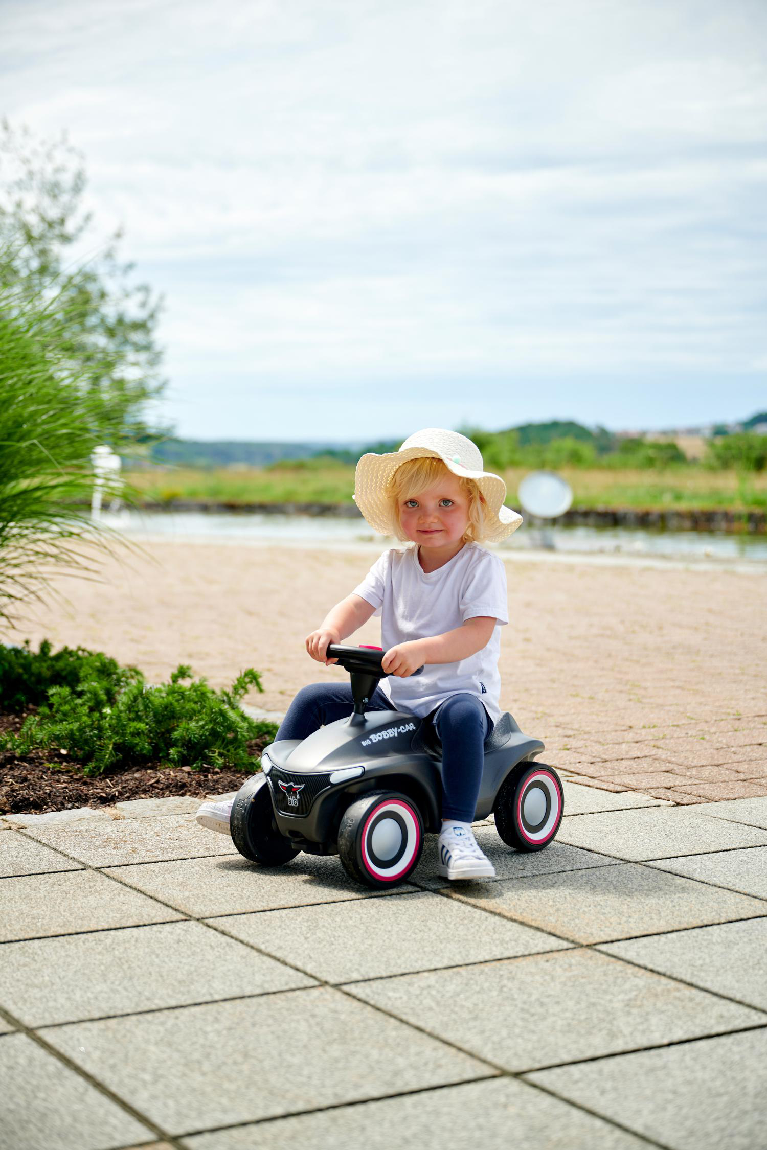 Ein junges Kind mit Strohhut sitzt draußen auf einem Spielzeugauto. Das Auto ist grau mit roten Rädern. Hintergrund hat Wasser und Grün.