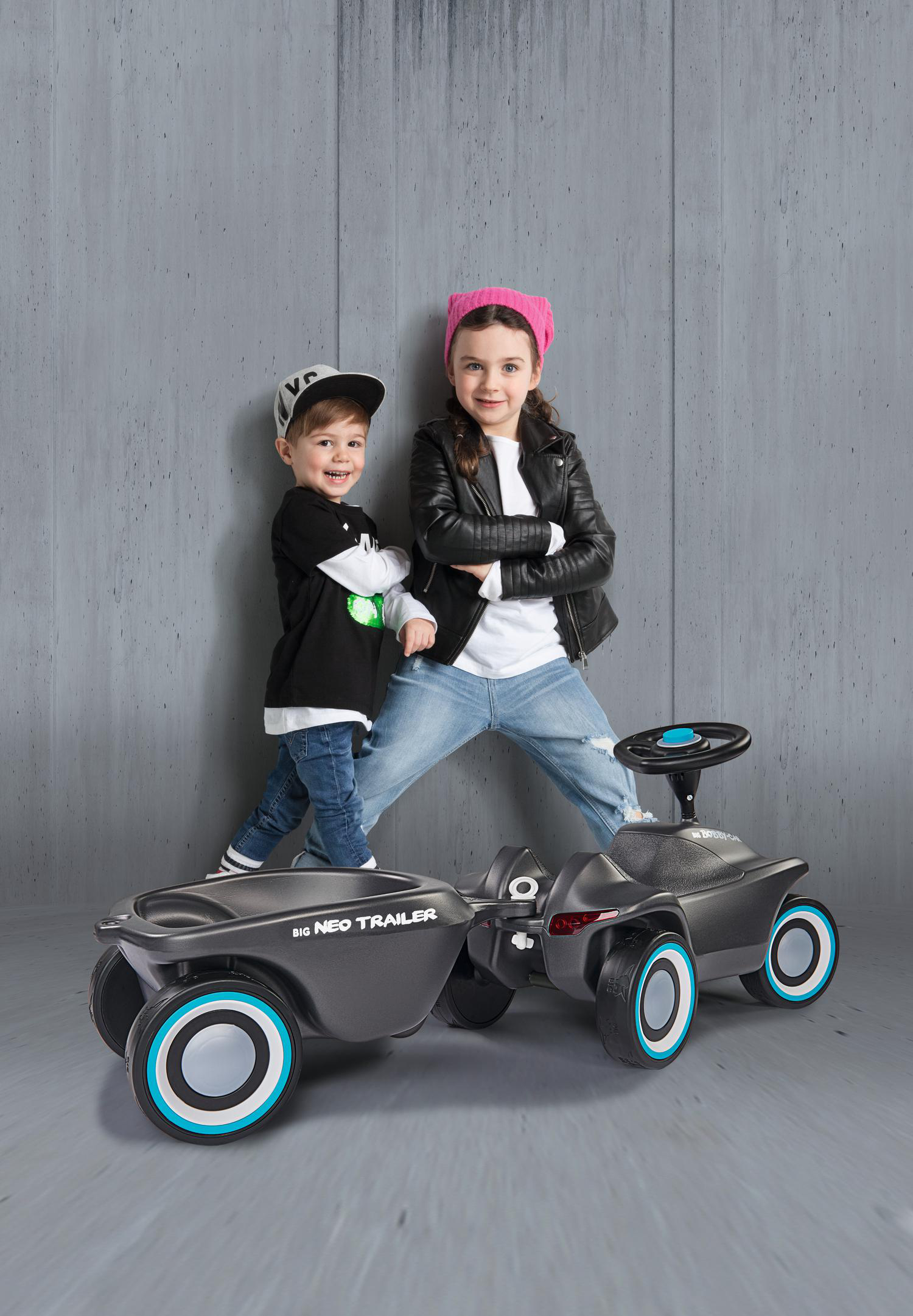 Zwei Kinder stehen neben einem Spielzeugauto und Anhänger. Eines trägt einen rosa Hut und eine schwarze Jacke. Das andere trägt eine Kappe und ein schwarzes Hemd.