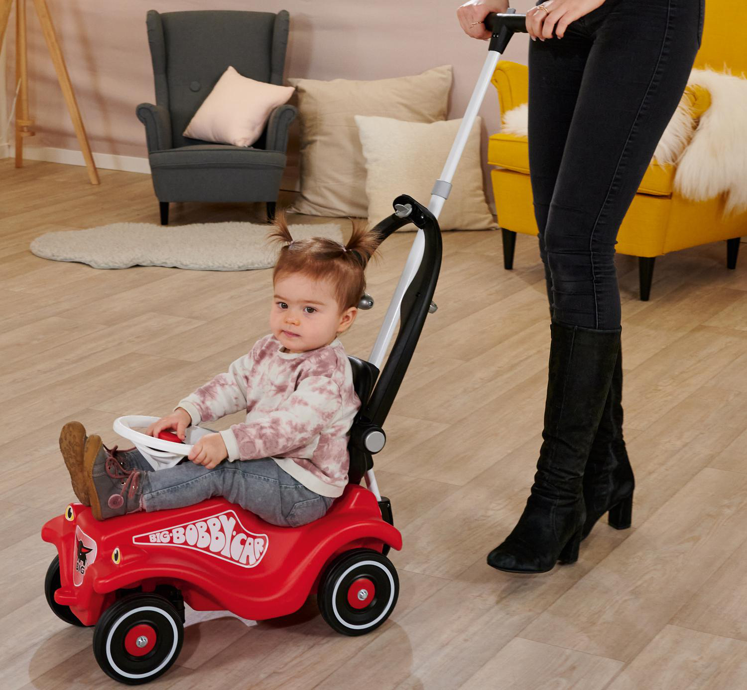 Ein Baby in einem roten Spielzeugauto wird von einer Frau geschoben. Die Szene umfasst einen grauen Sessel, ein gelbes Sofa und Holzböden.