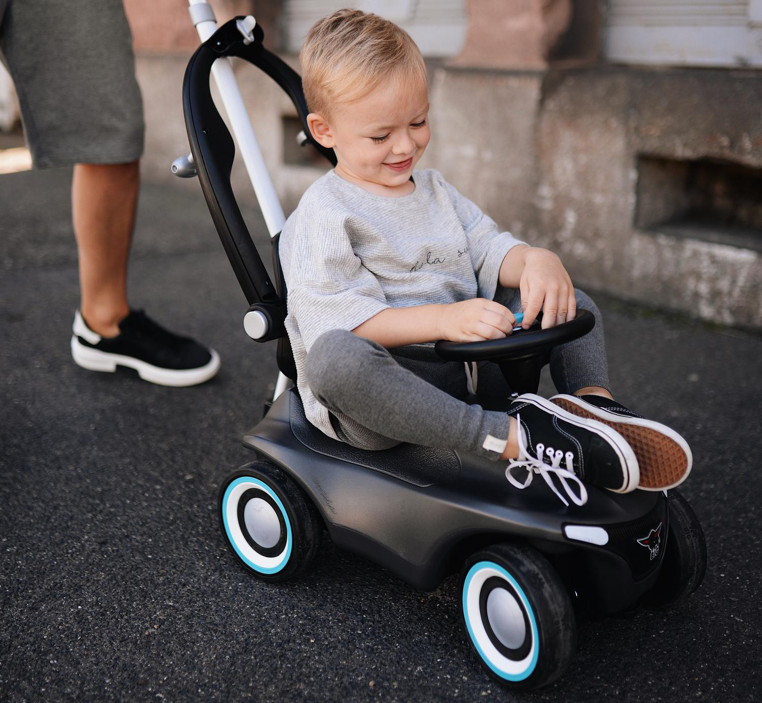 Ein Kleinkind sitzt lächelnd in einem schwarzen Spielzeugauto. Ein Erwachsener schiebt das Auto. Das Auto hat weiße und blaue Details.