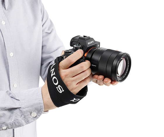 Une personne tient un appareil photo Sony noir avec un objectif. L'appareil photo a une dragonne et la personne porte une chemise bleu clair.