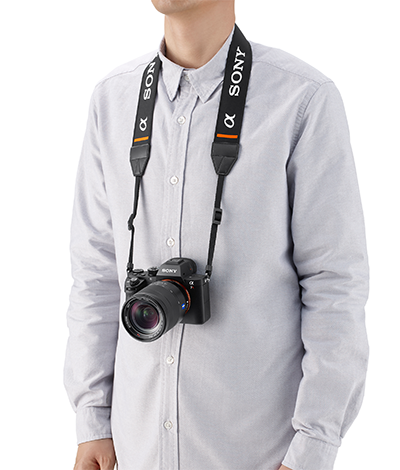 Un homme porte un appareil photo Sony sur une sangle. L'appareil photo est suspendu devant sa poitrine. Il porte une chemise gris clair.