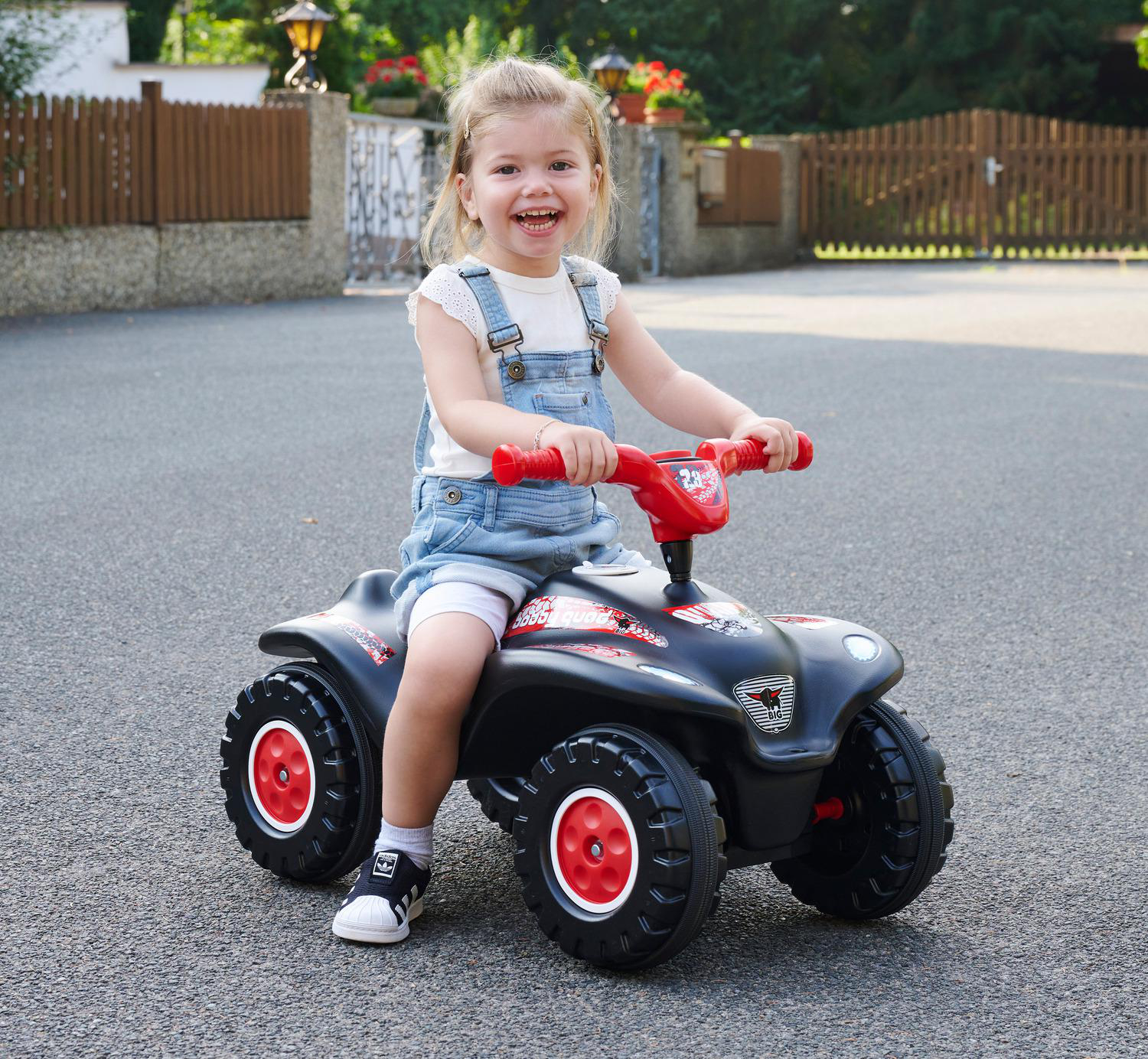 Ein lächelndes Mädchen in Latzhosen sitzt auf einem schwarzen Spielzeug-ATV mit roten Akzenten. Sie ist auf einer asphaltierten Straße, mit einem Zaun und Grün im Hintergrund.
