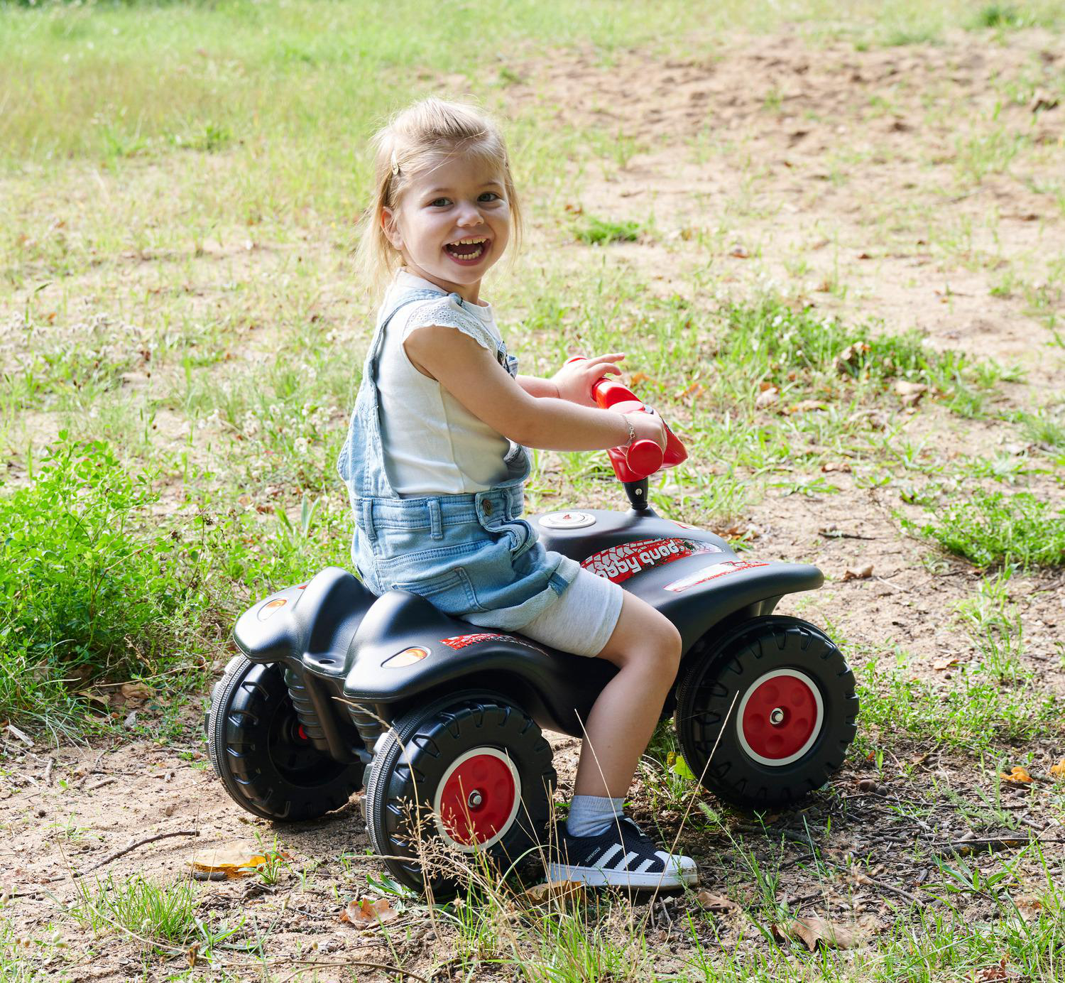 Ein junges Mädchen lächelt, während es auf einem schwarzen und roten Spielzeug-ATV sitzt. Sie trägt Jeans-Overalls und Turnschuhe in einer grünen Gegend.