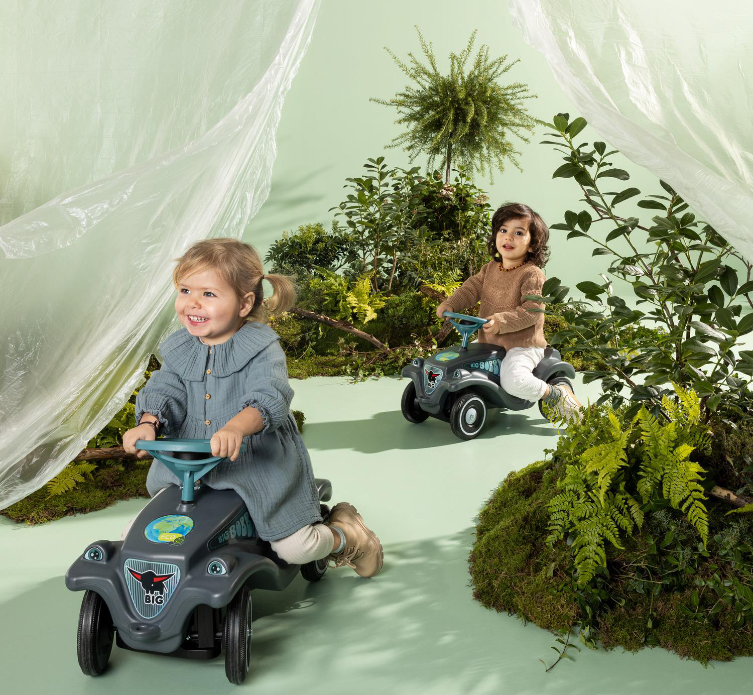 Zwei Kinder fahren in einer grünen Umgebung mit Pflanzen und einem durchscheinenden Hintergrund mit grauen Spielzeugautos.