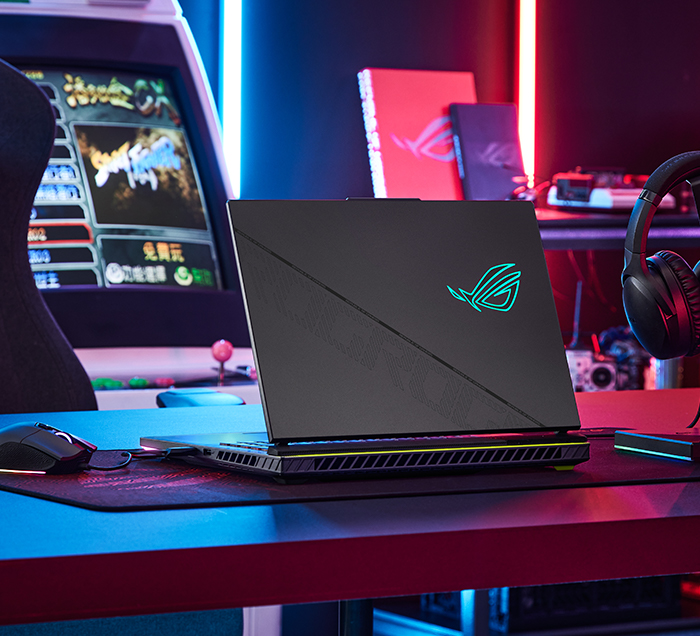 Ein schwarzer Laptop mit leuchtendem Logo steht auf einem Schreibtisch. Im Hintergrund sind ein Spielautomat und Kopfhörer zu sehen, mit roten und blauen Lichtern.