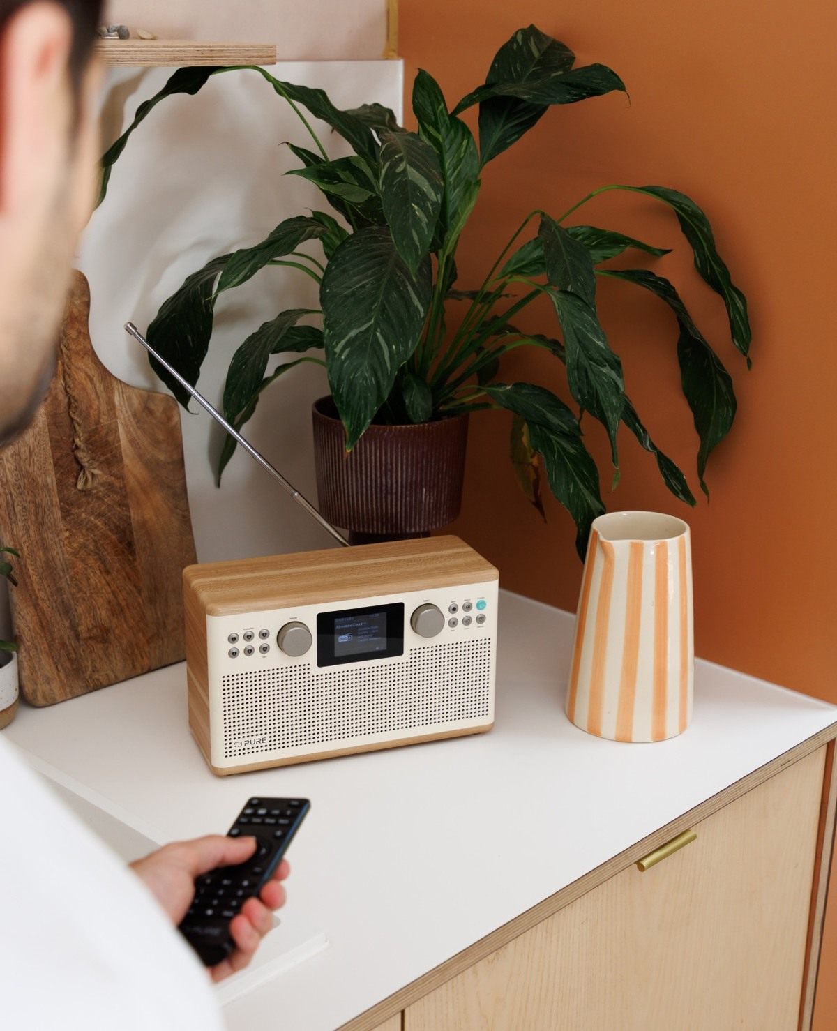 Une personne utilise une télécommande pour contrôler une radio sur une surface blanche, avec une plante et un vase en arrière-plan.