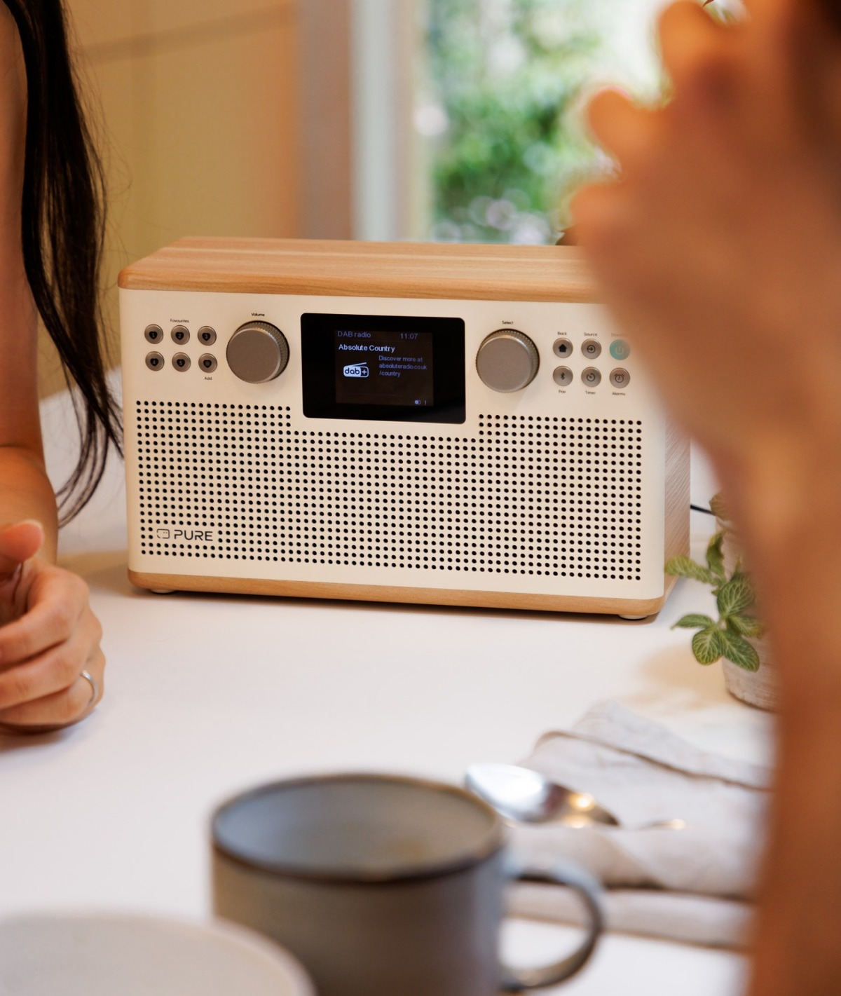Une radio en bois sur une table blanche. Deux personnes sont proches. Une tasse et une cuillère au premier plan. Une petite plante est visible.