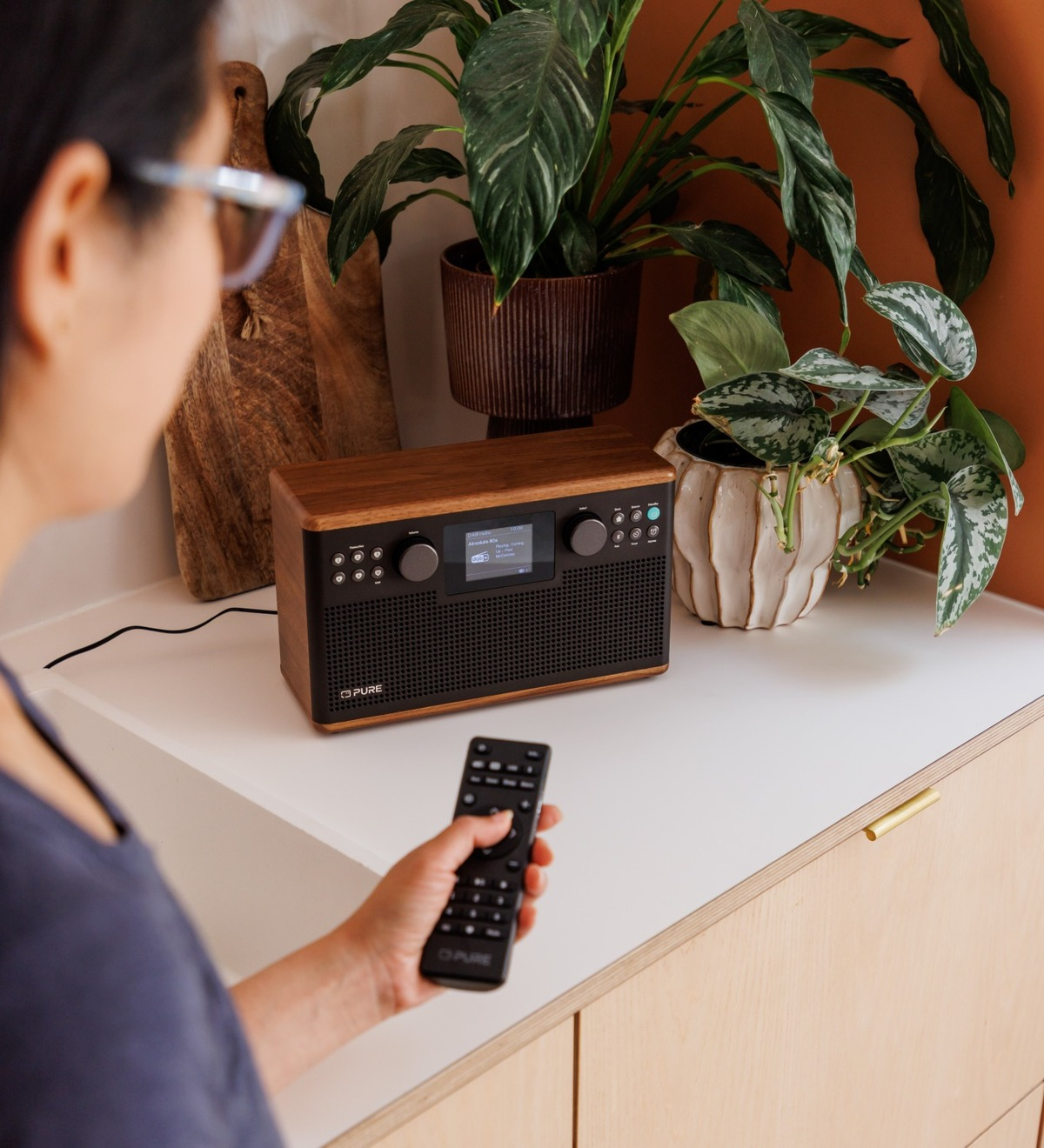 Une personne utilise une télécommande pour une radio en bois sur une surface blanche, avec des plantes en arrière-plan.