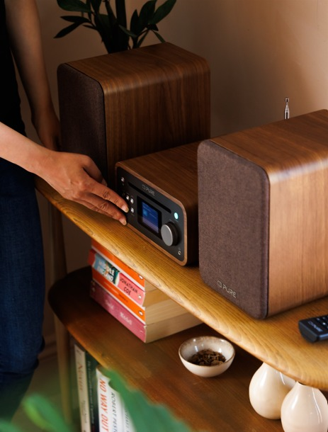 Una mano gira una manopola su uno stereo in legno. Libri e piccole ciotole sono sullo scaffale sottostante. Gli altoparlanti affiancano lo stereo.