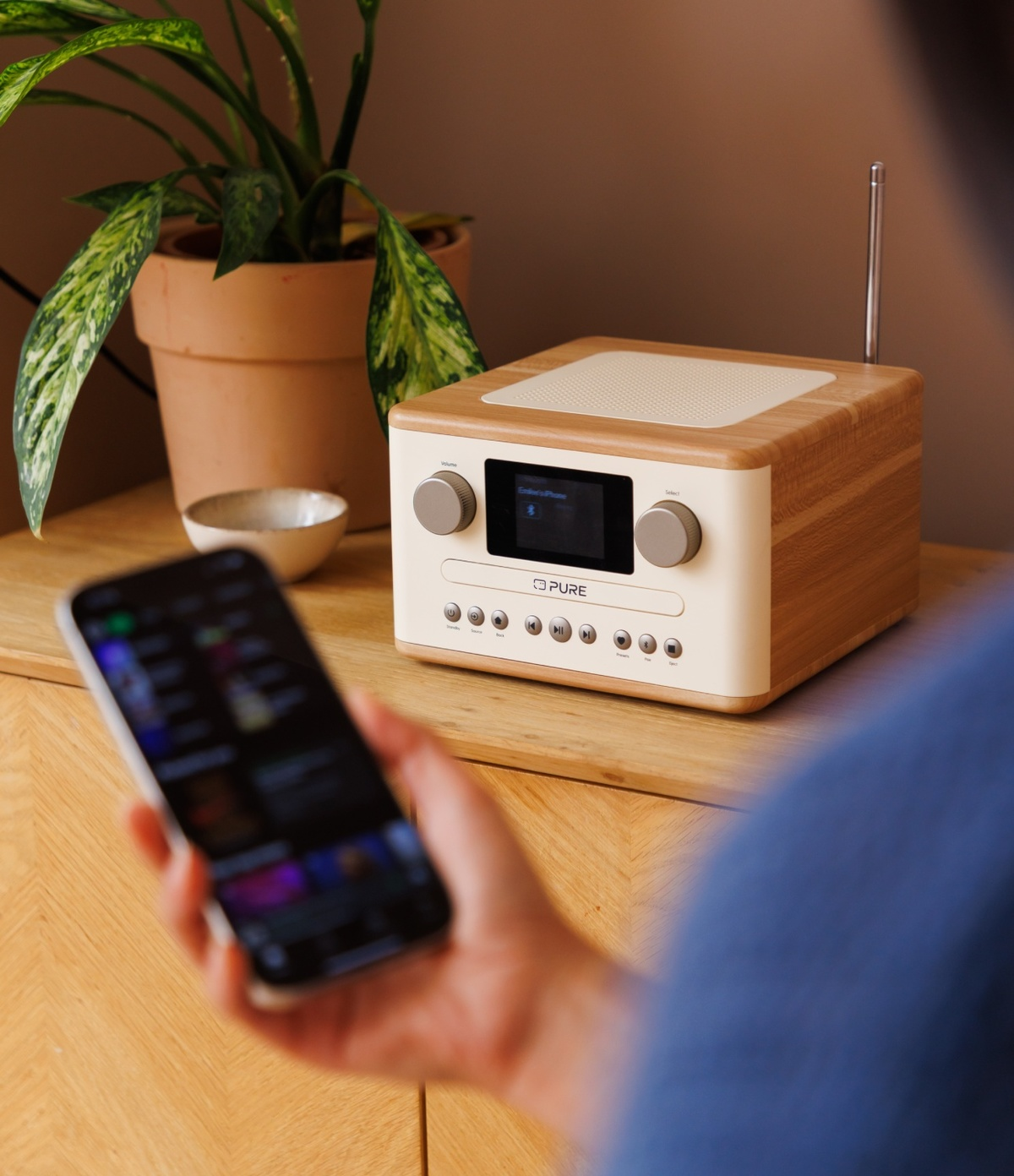 Une personne tenant un téléphone près d'une radio sur une table en bois. Une plante et un petit bol en arrière-plan.