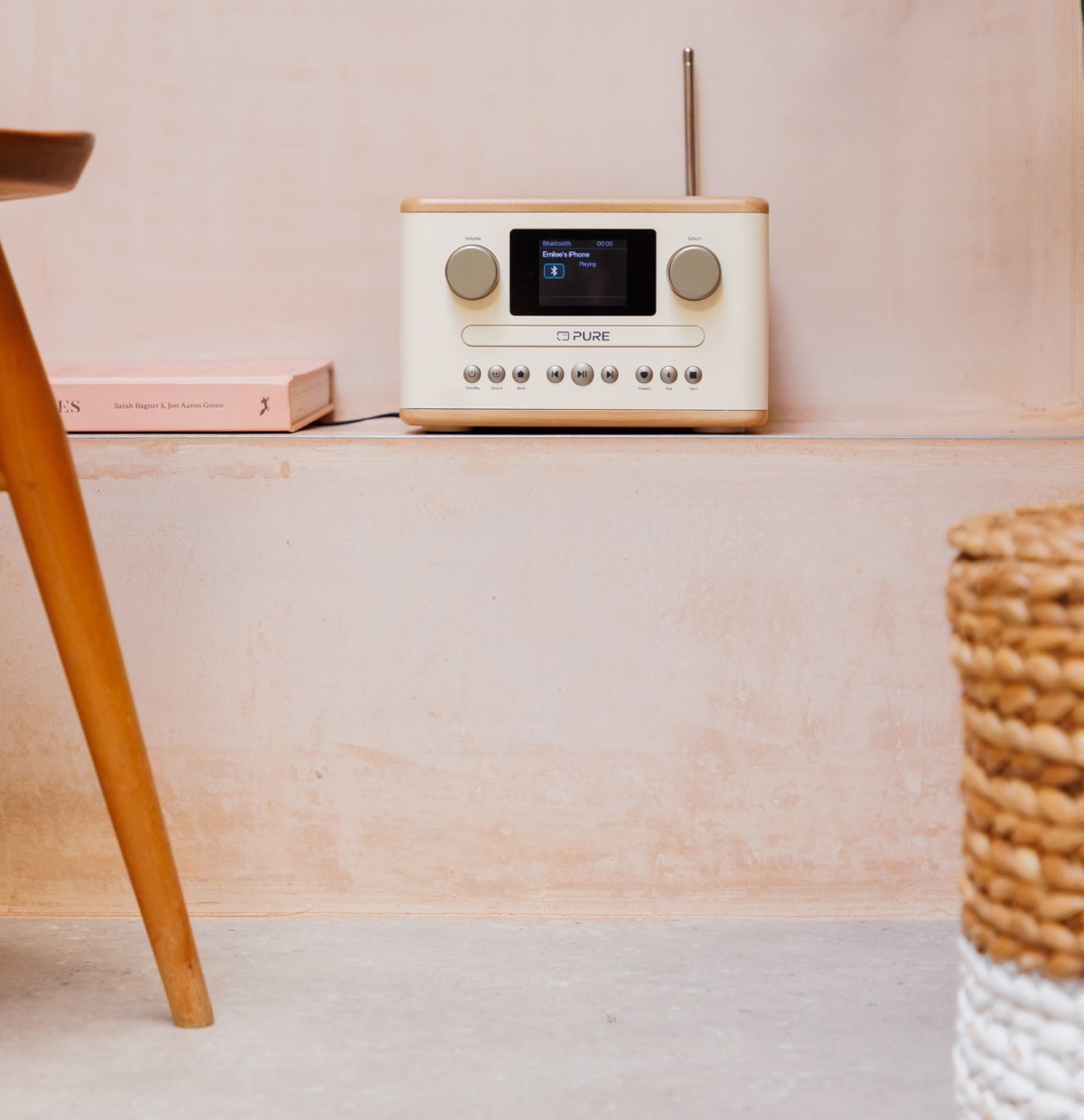 Radio crème avec des accents en bois sur une étagère. Un livre et un pied de table à gauche. Un panier en osier à droite.