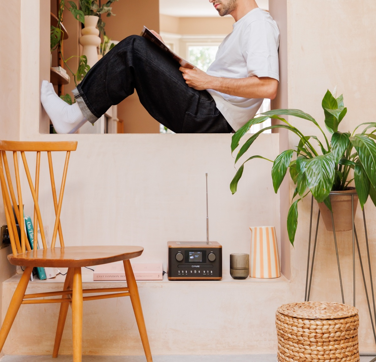 Un homme lit dans une fenêtre. Chaise, radio, plantes et un panier en osier sont dans la pièce. Les couleurs sont beige et marron.